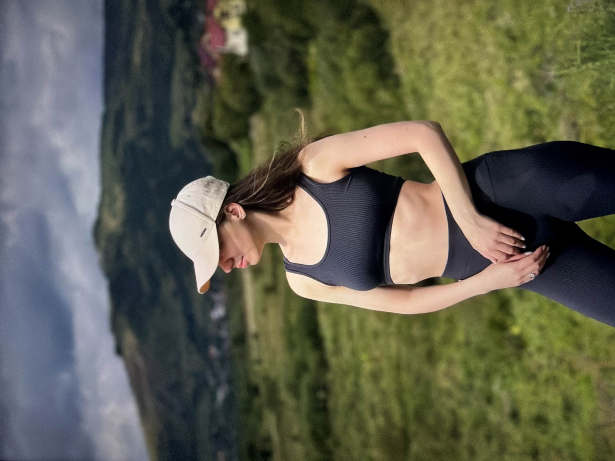 A woman wearing a black sports bra and leggings, and a light beige baseball cap is standing in a grassy field with hills and some houses in the background. She is facing left and looking away from the camera. Her hands are clasped in front of her waist. The sky above is cloudy and dark. The location is identified as Unknown, Unknown.