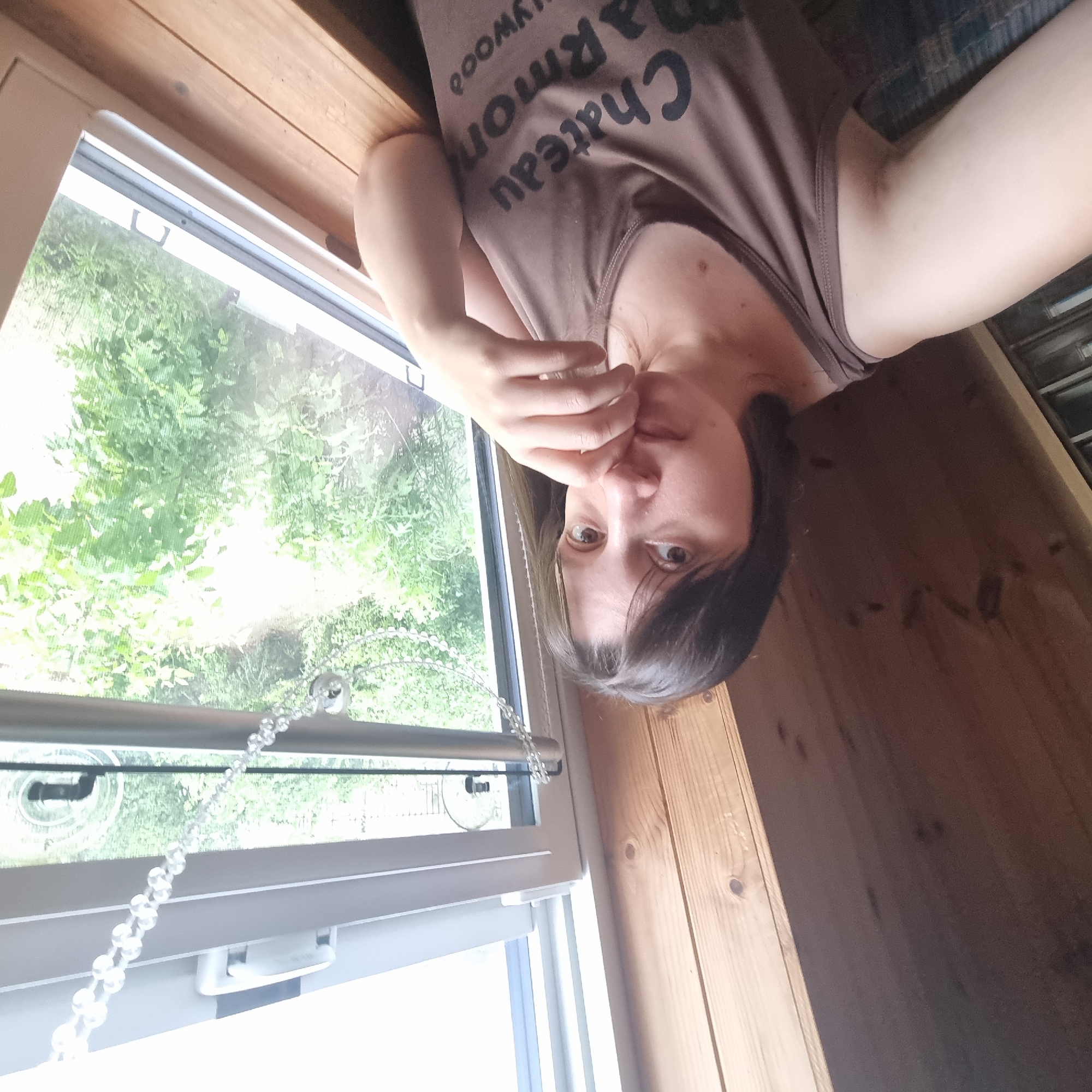 A selfie taken by a woman with dark hair and brown eyes, partially obscuring her face with her hand. She is wearing a brown tank top with the text "Chateau Marmon Hollywood" printed on it. The woman is positioned indoors, leaning against a wooden surface, with a window to her right showing lush green foliage outside. The wooden interior walls are visible to her left. The scene appears to be in a room with natural light filtering in from the window. The location is not specified.