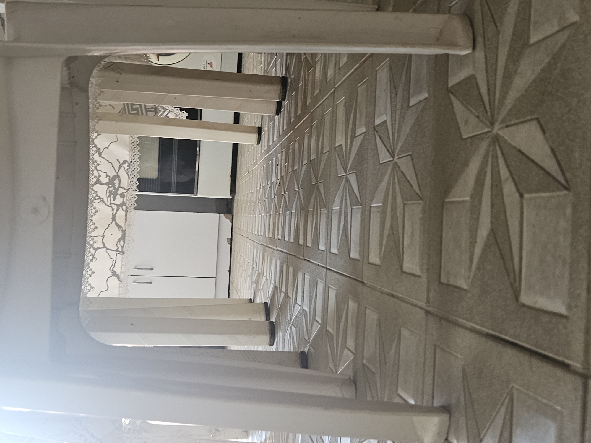 The image is taken from a low angle, looking through the legs of white plastic chairs. The chairs are arranged on a tiled floor with a geometric pattern. In the background, there is a white cabinet with silver handles and a black appliance, possibly a stove or oven, partially visible. A lace curtain hangs across the gap between the chairs, obscuring some of the background. The lighting is bright and appears to be natural light from the left, creating strong shadows from the chair legs on the floor. There are no people or animals visible in the image. The scene suggests an indoor setting, possibly a kitchen or dining area.