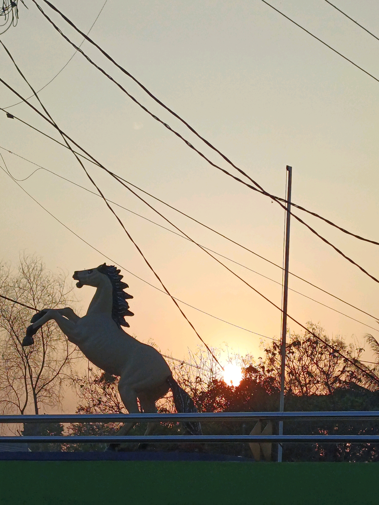 A statue of a rearing horse is silhouetted against an orange sky at sunset in Krimun, Indonesia. The statue is a pale color and appears to be made of concrete or stone. It is positioned on a raised platform, with a metal railing in front of it. In the background, there are silhouetted trees and a cluster of power lines crisscrossing the sky. The sun is partially obscured by the trees, casting a warm glow. The ground in the foreground is a dark green.
