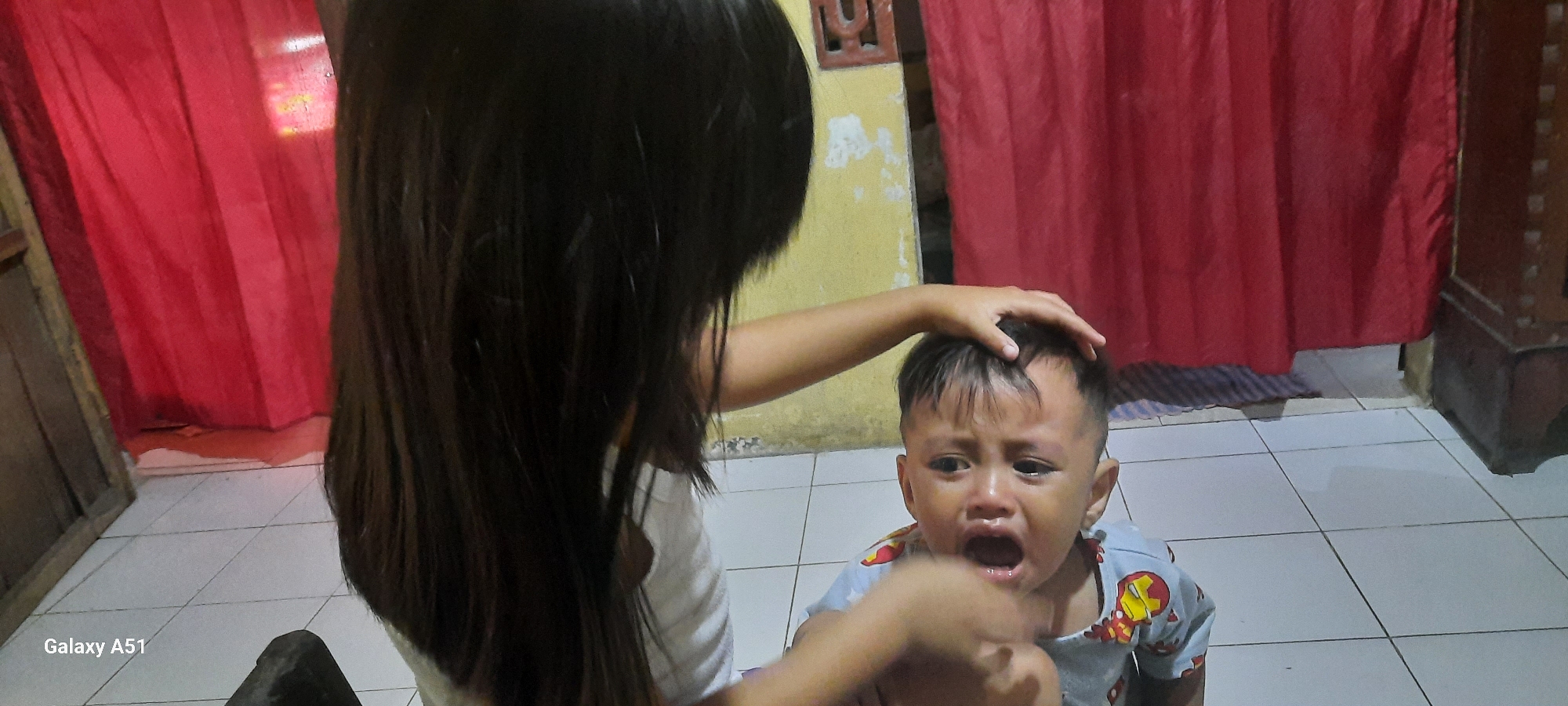 In an indoor setting, likely a room in a home in Ploso, Indonesia, a young child is crying intensely. The child, appearing to be a toddler, is wearing a light blue shirt with a repeating cartoon character pattern, possibly Iron Man. The child's face is red with tears streaming down, eyes wide open, and mouth agape. An older child, possibly a girl with long dark hair, is visible from behind, with one hand touching the crying child's head, perhaps attempting to comfort or check on them. The background features red curtains, a yellow wall, and a tiled floor. To the left, a wooden door frame is visible, and to the right, a dark wooden piece of furniture is present. The overall impression is of a domestic scene where a child is experiencing distress.