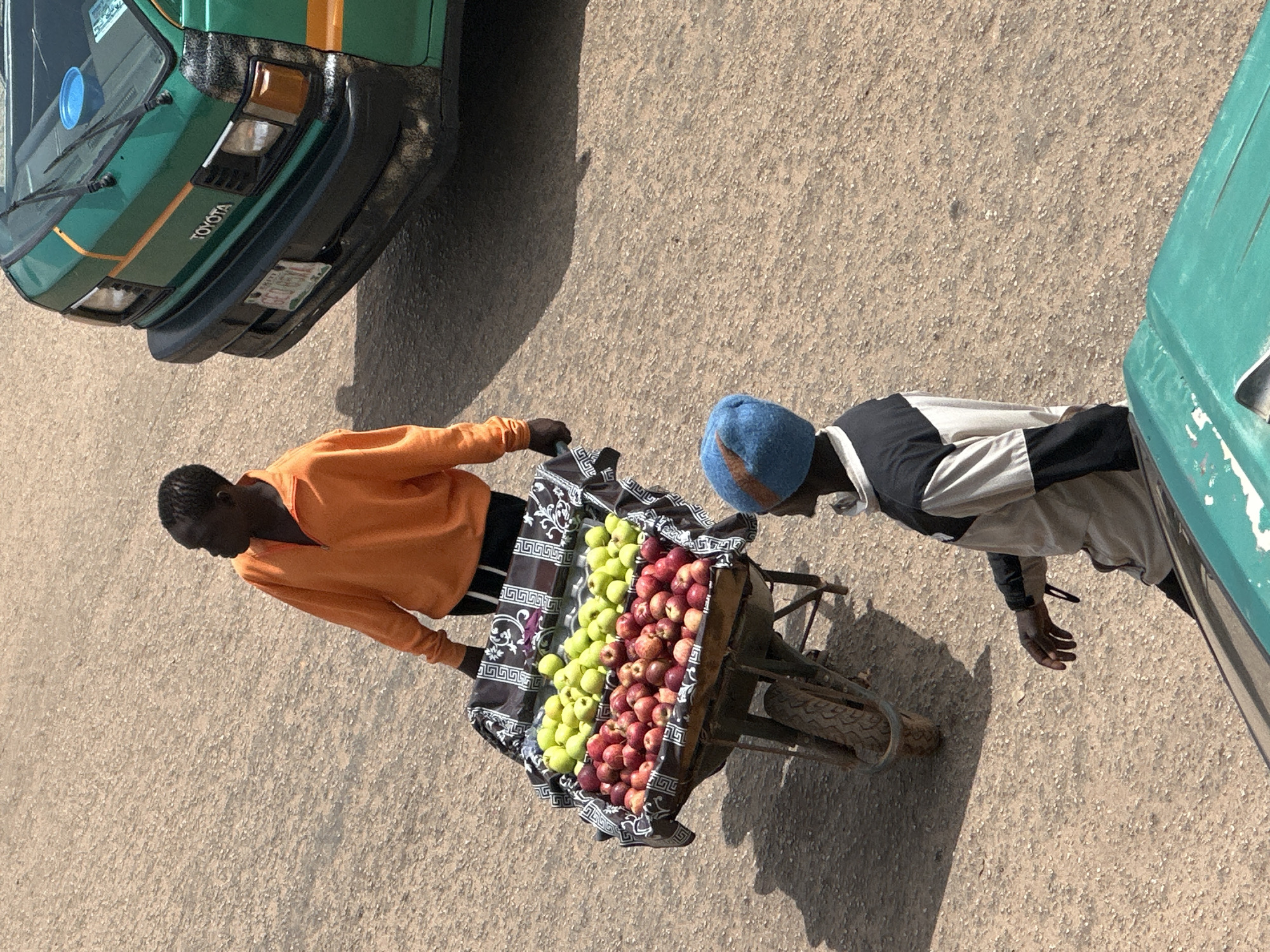 In Bukuru, Nigeria, a fruit vendor in an orange sweatshirt pushes a wheelbarrow laden with apples and other fruits. He is walking alongside a green Toyota van. Another person wearing a blue beanie and a grey and black jacket is standing nearby. The ground is a dusty, light brown surface, possibly a road or unpaved area. The scene is lit by bright sunlight, casting distinct shadows. The wheelbarrow is decorated with a black and white patterned fabric. The van has a license plate that reads "GEO HB4."