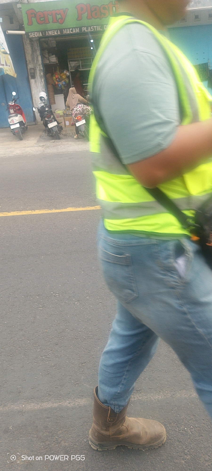 A person walks on a paved road, wearing light blue jeans, a light teal t-shirt, and a bright yellow safety vest with reflective gray stripes. They are also wearing brown work boots. In the background, there is a building with a green sign that reads "Ferry Plastik". Several motorcycles are parked in front of the building, and two people are visible inside the store. The scene appears to be in a commercial area, possibly a street in a city.