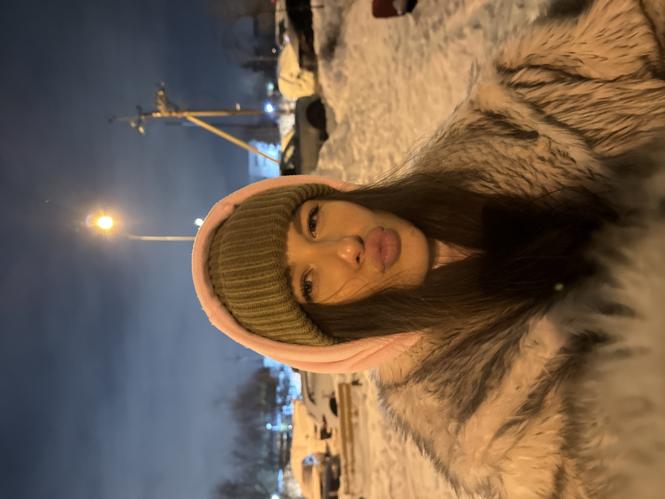 A young woman poses outdoors at night. She wears a knit cap with a pink hood and a large fur-like coat. Snow covers the ground in the background, with blurred lights and shapes of parked cars visible. Overhead lights illuminate the scene. The woman has long dark hair and her face is the focal point, capturing her expression with pursed lips.