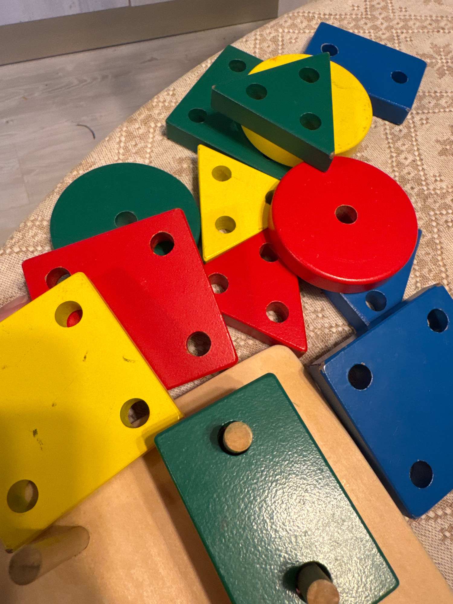 The image displays a collection of colorful wooden geometric shapes scattered on a textured, light-colored fabric. The shapes include red and yellow squares, a green circle, a green triangle with yellow and red sections, and blue rectangular blocks. Several shapes have circular holes drilled through them. The lighting suggests an indoor setting, possibly a home or classroom. There are no people visible in the image. The scene does not provide any information about the city of Saint Petersburg, Russia, or any specific location within it.