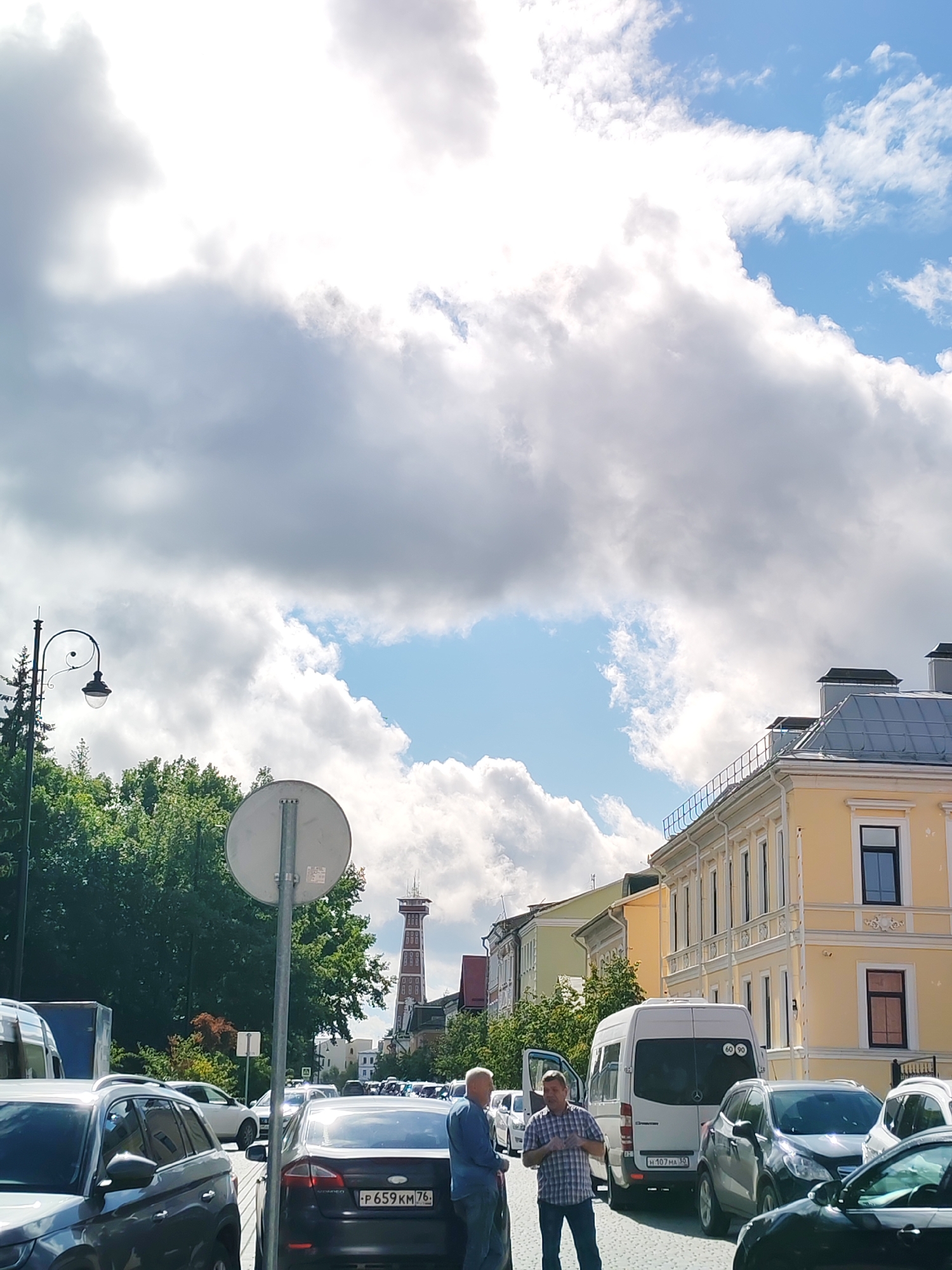 This photograph captures a street scene in Bol'shie Mkhi, Russia, under a dynamic sky filled with both bright sunlight and heavy, dramatic clouds. The time of day appears to be late morning or early afternoon, suggested by the strong light and shadows.
In the foreground, several vehicles are parked or in motion on a cobblestone street. A dark SUV is on the left, and a dark sedan with a visible license plate "P 659 KM 76" is in the center. Further back, a white van is parked, and two men are standing near its open side door, engaged in conversation. One man is wearing a blue plaid shirt and dark pants, while the other is in a blue shirt. A few other cars are visible in the background, suggesting a moderately busy street.
The middle ground features lush green trees lining the street, creating a verdant backdrop. A classic street lamp with a curved arm stands tall on the left. Beyond the trees, a distinctive red and white tower with a grid-like structure rises above the buildings, possibly a communication tower or historical landmark.
The architecture visible includes a prominent yellow building with multiple windows and ornate trim on the right, and other buildings with light-colored facades further down the street. The sky is a prominent element, with a large patch of bright blue sky visible through the clouds, which are illuminated by the sun, creating areas of intense brightness and dark shadows. The overall impression is a partly cloudy day with dynamic lighting.