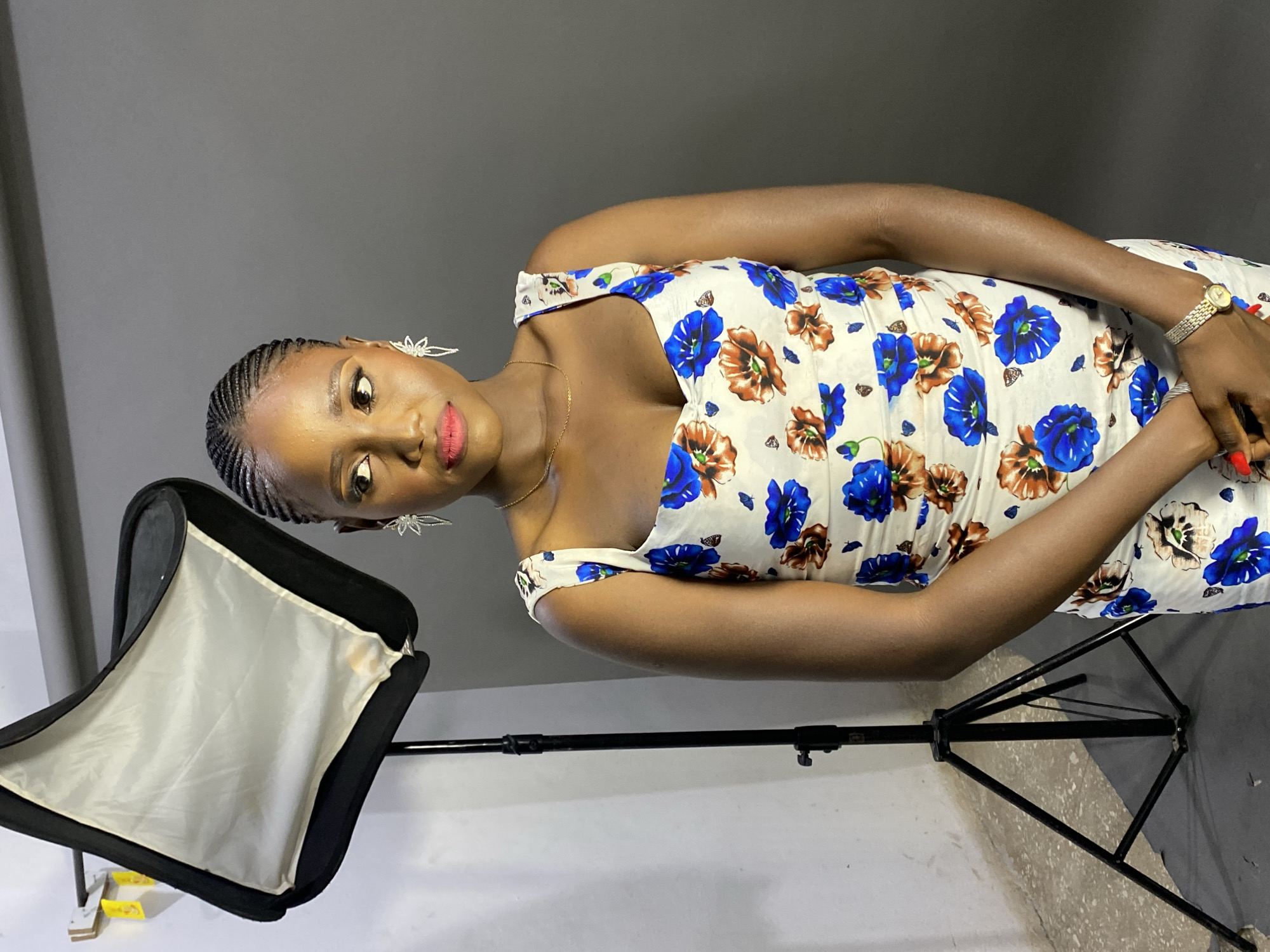 A young woman is featured in a studio portrait, likely taken in Jos, Nigeria. She has dark skin and her hair is styled in cornrows pulled back from her face. She looks directly at the camera with a confident gaze and a slight smile, wearing full makeup including red lipstick and winged eyeliner.
She is dressed in a white, off-shoulder dress featuring distinct blue and brown floral patterns. Accessories include delicate, star-shaped or abstract white earrings, a thin gold chain necklace, and a gold-toned watch on her left wrist. Her fingernails are painted red.
The setting appears to be an indoor studio with a plain, solid gray background and a lighter gray floor or lower wall. To the woman's left, a black softbox on a stand is visible, indicating artificial lighting used for the photoshoot. No legible text is discernible in the image.
