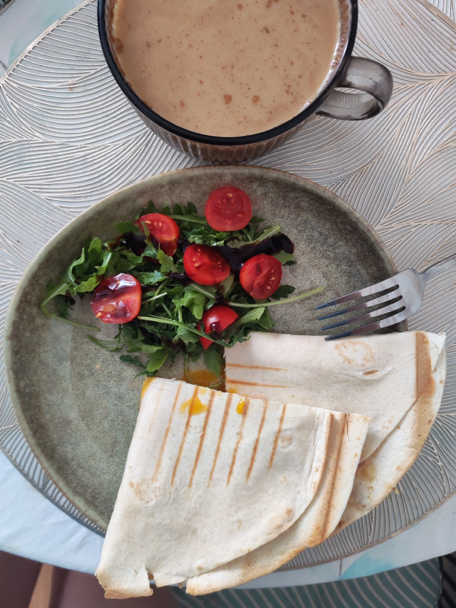 A bird's-eye view shot shows a meal on a textured placemat. In the upper portion of the image, a clear glass mug filled with a light brown beverage, possibly coffee with milk, is visible. Below the mug, a round, grey ceramic plate holds a salad of arugula and halved cherry tomatoes, drizzled with a dark sauce. Beside the salad are two folded quesadillas, showing grill marks and a yellow filling. A silver fork rests on the plate, positioned between the salad and the quesadillas. The background consists of a light-colored placemat with a wave-like pattern. No people are present in the scene. The overall impression is that of a breakfast or light meal. This scene is not specifically identifiable to a particular location in Izhevsk, Russia, or any other city without further contextual information.