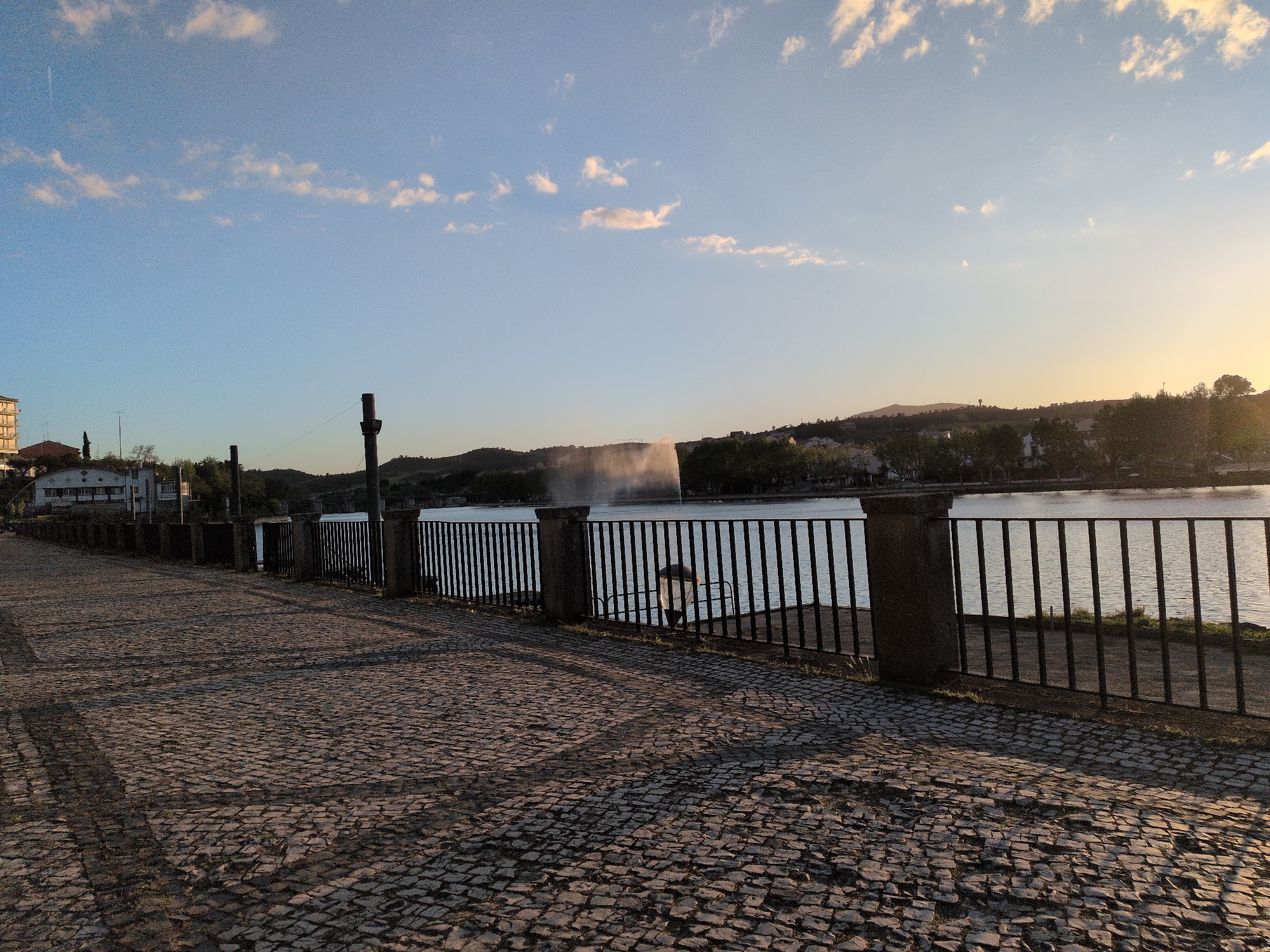 A view along a cobblestone pathway by a body of water, potentially the Tua River, in Mirandela, Portugal. The path is bordered by a dark iron fence with stone posts. In the water, there's a fountain spraying water upwards. Across the water, there are buildings and hillsides, with some trees. The sky is light blue with scattered white clouds and the sun is setting, casting a warm glow. The image is taken at ground level, with a wide view of the scene.