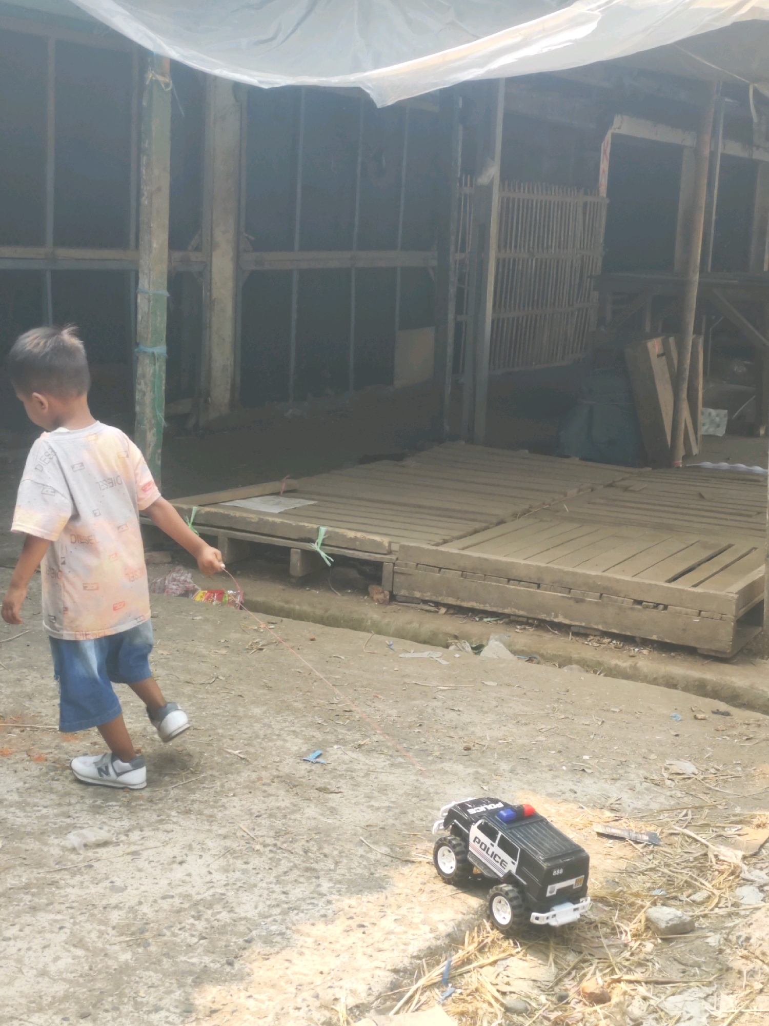 A young boy, with short dark hair, wearing a white t-shirt with colorful graphics and blue denim shorts, walks away from the camera, pulling a red string attached to what appears to be a toy. The boy is wearing white sneakers. Beside him on the ground, to the right, is a black toy police car with white detailing, large tires, and flashing blue and red lights. The background features an open-air structure with wooden planks laid out on the ground and vertical supports, possibly indicating a market or construction area. The ground is a mix of concrete and dirt, with some scattered debris. The scene is outdoors and appears to be a sunny day, with sunlight casting shadows. The location is identified as Karawang, Indonesia.