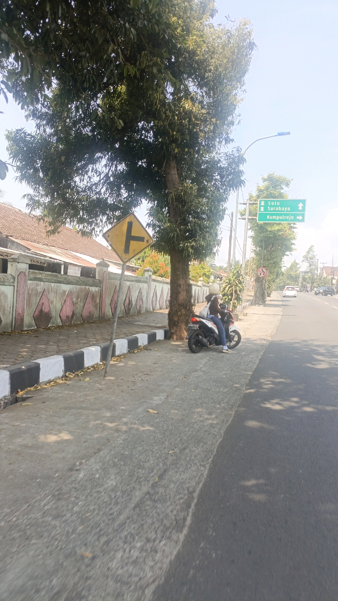 A woman wearing a blue shirt and jeans is sitting on a motorcycle parked on the side of a road in Salatiga, Indonesia. The motorcycle is white and black. She is looking towards the left side of the image. In the background, there is a wall with decorative patterns and trees. A yellow diamond-shaped traffic sign with a black "T" intersection symbol is also visible near the motorcycle. Further down the road, there is a green directional sign indicating "Solo" with an upward arrow, "Surabaya" with an upward arrow, and "Kumpulrejo" with a right arrow. The sky is clear and blue. The road is paved and appears to be an urban or suburban area.