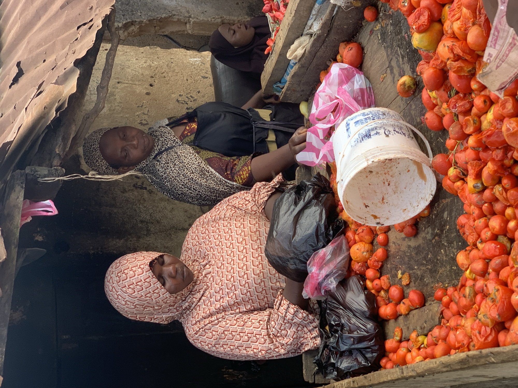 In Jos, Nigeria, three women are lying down on a wooden surface, which is surrounded by a large quantity of tomatoes, some of which appear rotten. One woman is wearing a patterned headscarf and outfit with black plastic bags nearby. Another woman is wearing a leopard-print shawl and a black dress. The third woman is wearing a dark-colored head covering. A white bucket and plastic bags are also present. The scene is enclosed by weathered structures.
