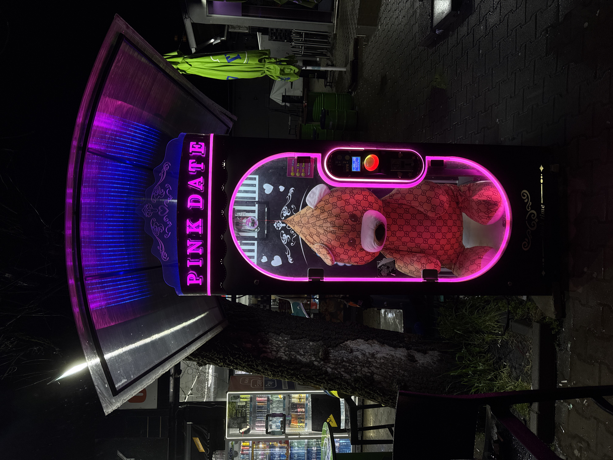 The image depicts a "Pink Date" claw machine in Sofia, Bulgaria, at night. The machine is brightly lit with pink and blue neon lights, showcasing a teddy bear inside the claw area. A beverage vending machine is visible next to it. The surroundings appear to be an outdoor urban setting, with wet pavement and some street furniture.