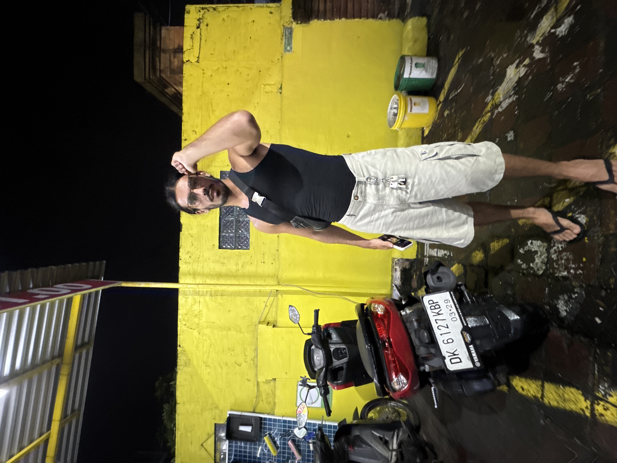 A man stands beside a motorbike in North Kuta, Indonesia. The motorbike's license plate reads "DK 6127 KBP" and has a date of "03-29". The man is wearing a black tank top, light-colored shorts, and sandals. He is holding a phone and appears to be adjusting his hair. The background includes a yellow building and an awning. The ground is wet.