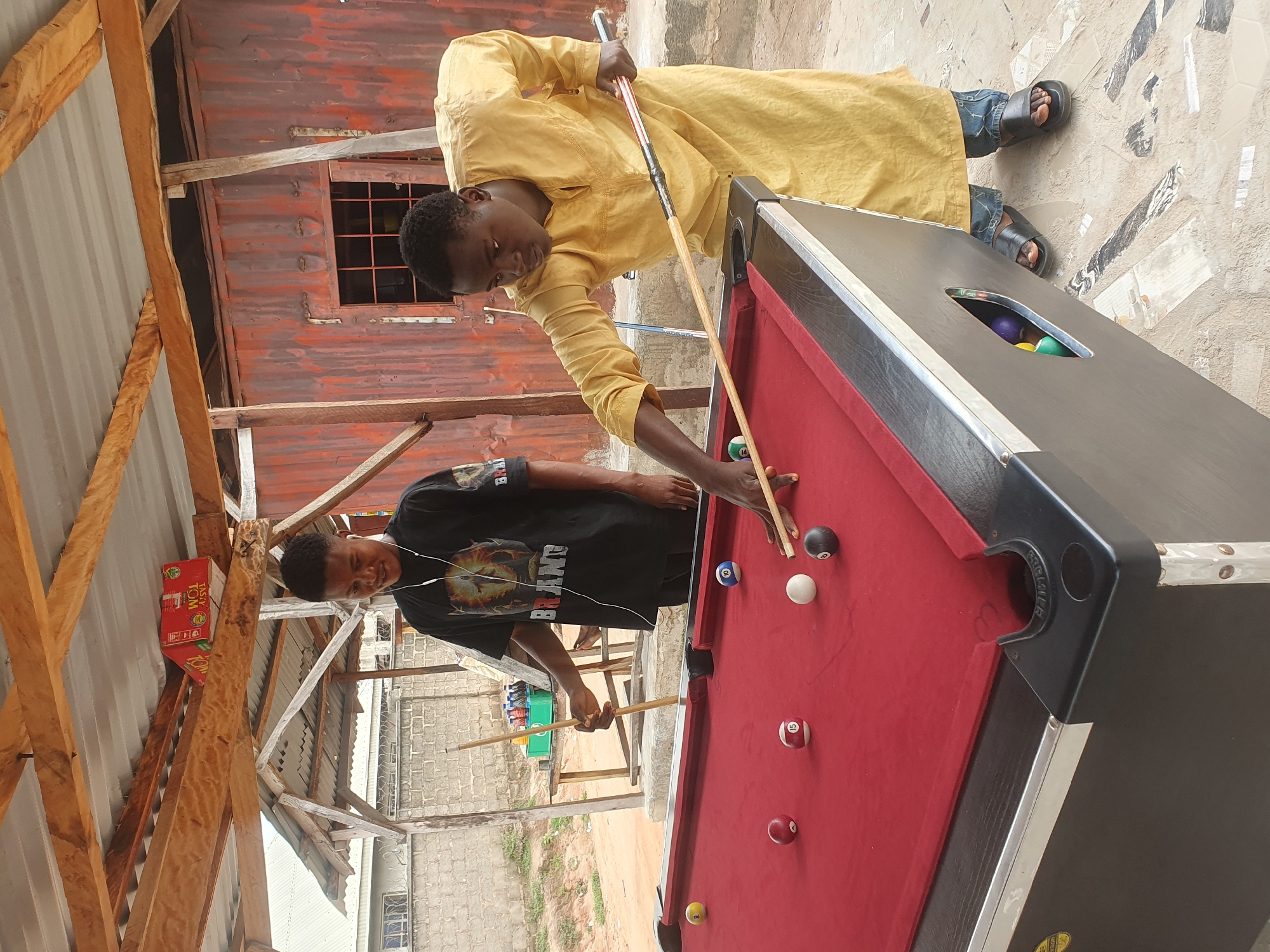In Lokoja, Nigeria, two young men are engaged in a game of pool. One man, wearing a black t-shirt with graphics and white earbuds, stands on the left, holding a pool cue. The other man, dressed in a yellow robe and blue jeans, is on the right, actively playing, aiming his cue at a billiard ball. The pool table has a red felt surface with several colored balls scattered across it, including a yellow ball near the left pocket, a red ball and a striped ball numbered 15 to its right, a white ball and a black ball numbered 8 further to the right, and a striped ball numbered 10 near the right side. In the background, a corrugated metal structure with a wooden roof suggests an outdoor or semi-outdoor setting, with some crates visible on a shelf above. The ground is paved with patterned tiles and concrete. The overall scene depicts a casual leisure activity in a Nigerian urban environment.