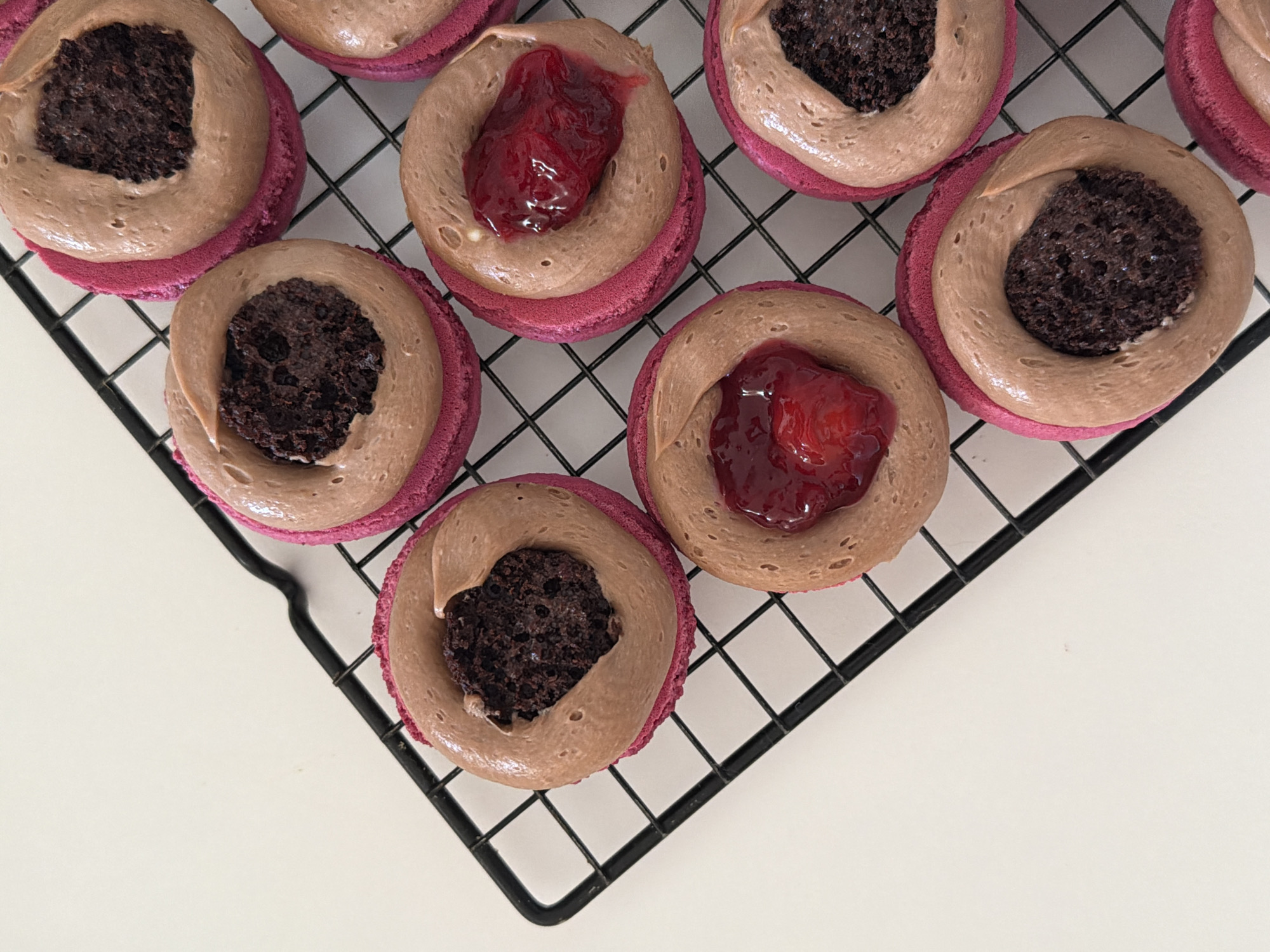 The image shows several macarons arranged on a black wire cooling rack, which sits on a white surface. The macarons have a pink shell and are topped with a circular ring of light brown frosting. Some of the macarons are filled with a dark, textured substance that resembles brownie crumbles, while others are filled with a red, jelly-like substance. The macarons are positioned in a partial triangular formation within the frame. The scene is brightly lit, with a focus on the details of the pastries. There is no indication of the location being Tel-Aviv, Israel.