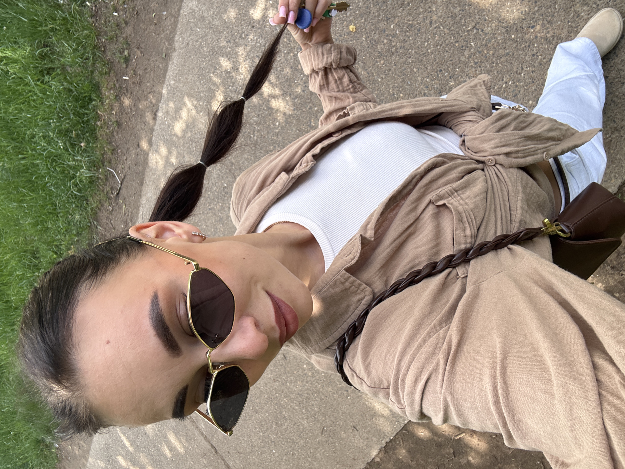 A young woman stands outdoors in Chaikovsky, Russia. She wears gold-rimmed sunglasses, a white ribbed tank top, a tan shirt tied at the waist, white pants, and beige shoes. She has a brown shoulder bag with a braided strap. Her dark hair is styled in segmented ponytails secured with hair ties. In her raised hand, she holds keys. She stands on a concrete surface next to a patch of grass. The photo appears to be taken at eye level, angled downwards towards the subject.