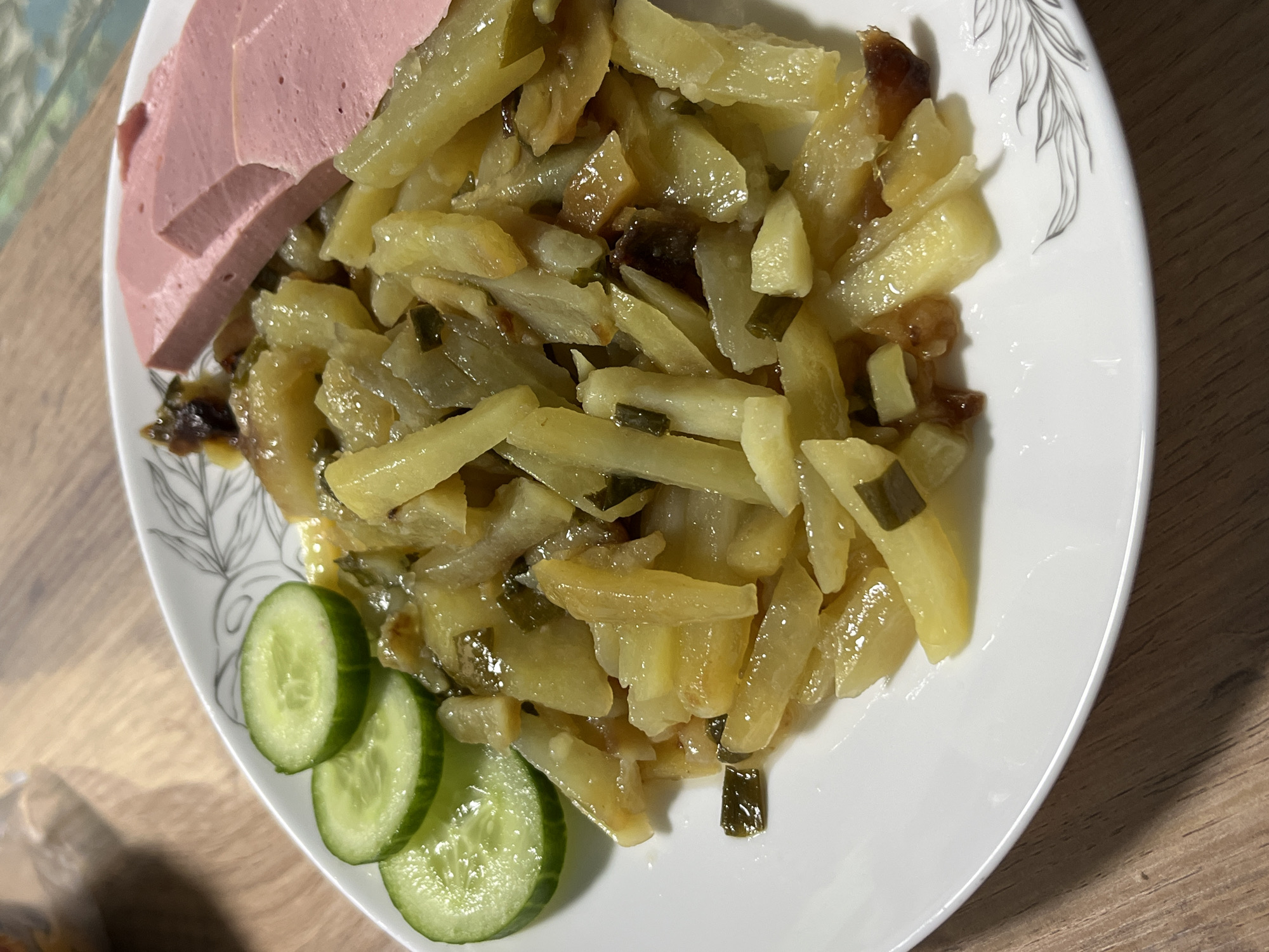 A plate of food sits on a wooden surface. The plate contains fried potatoes, sliced sausage, and sliced cucumbers. The plate is white with a grey leaf pattern on the edge. The location is likely in a home in Nizhny Tagil, Russia, given the typical ingredients and the cultural context. There are no people visible in the image.