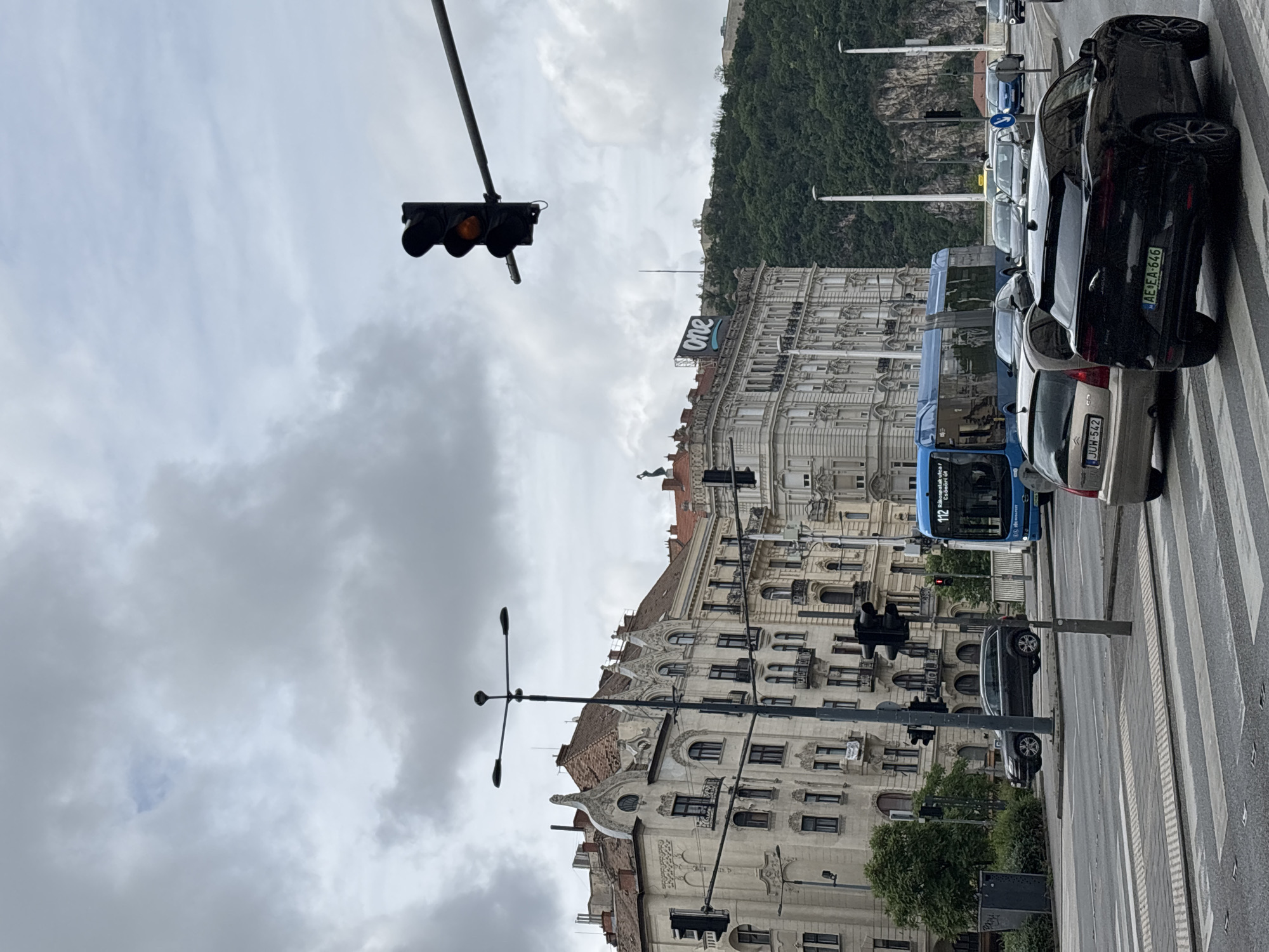 The image shows a street scene in Budapest, Hungary. The street is lined with buildings, including one with a prominent "one" logo on the roof. Traffic is present, with cars and a blue bus visible. A pedestrian crossing is marked on the road. Traffic lights are positioned along the street, one showing an orange light. In the background, a hill or mountain is covered in trees. The sky is overcast with clouds.