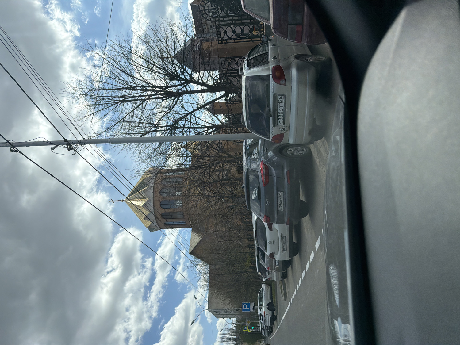 The image captures a street scene from the perspective of a vehicle, looking towards a row of parked cars and a distinctive building under a partly cloudy sky. The time of day is daytime, with bright light and scattered clouds allowing patches of blue sky to show through. Bare trees, indicating late fall, winter, or early spring, frame the background building.
In the midground, a line of cars is parked parallel to the road, which has white dashed markings. The closest visible car is a white hatchback with the Russian license plate "O330OT 126". Behind it is a gray SUV with the plate "E394OY 126". Further down the line, another white car displays "A069BH 126". All three visible license plates share the "126" region code, corresponding to Stavropol Krai, Russia, serving as a strong location cue. A partially obscured sign near one of the cars reads "EXIT". A blue parking sign with a white 'P' is visible further down the street, and a traffic light shows a green signal in the distance.
The background is dominated by a prominent stone or brick building, likely a church, characterized by its polygonal drum and a conical roof (cupola) topped with a cross. Utility poles and a network of overhead wires crisscross the sky, adding to the urban or semi-urban setting. The overall scene depicts a typical street environment with a mix of traffic infrastructure and religious architecture.