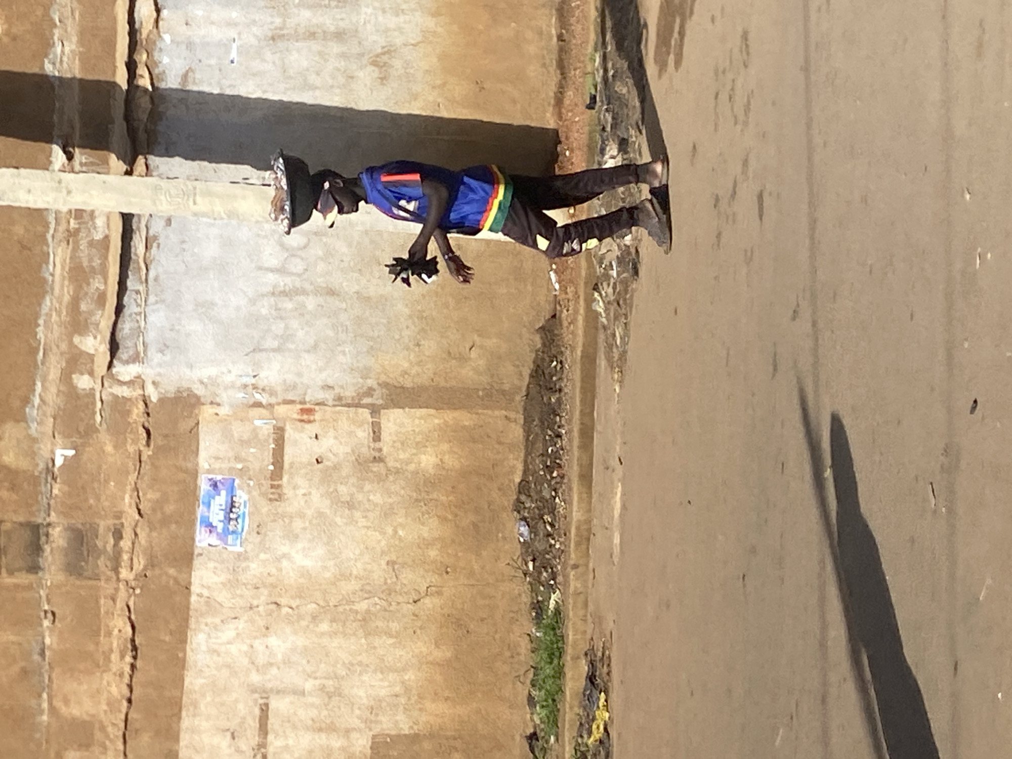 In Bukuru, Nigeria, a young man walks along a dirt road next to a weathered, light-colored wall. He is carrying a round, black bowl filled with what appears to be food balanced on his head. He wears a blue shirt with red and yellow trim on the sleeves and bottom, dark pants with yellow accents, a baseball cap, and sandals. In his left hand, he holds a small, dark object, possibly a toy. The ground is covered in debris, including trash and dry vegetation. A long shadow stretches across the road from the man and a nearby utility pole. The wall behind him is textured and shows signs of age and damage, with a small, faded poster affixed to it. The scene is illuminated by natural sunlight, casting distinct shadows.