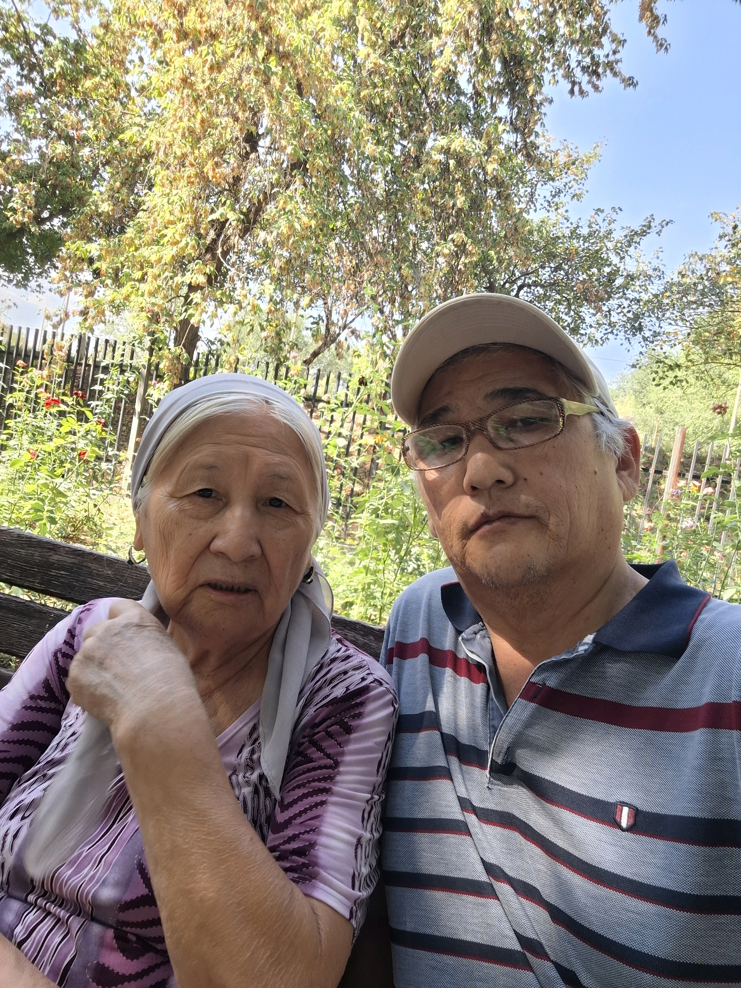 Two individuals, an older woman and a middle-aged man, are seated outdoors on a wooden bench, possibly in a garden or park setting. The woman, on the left, wears a light grey headscarf and a purple and white patterned shirt. She has earrings and her left arm rests on her lap. The man, on the right, wears a light beige baseball cap, glasses with gold-colored rims, and a blue, red, and grey striped polo shirt. They appear to be posing for a selfie. Behind them, a wooden fence is visible, along with rose bushes with red flowers and green foliage. A large tree with yellowing leaves dominates the upper portion of the image, with a clear blue sky visible through its branches. The overall impression is a candid, daytime photograph taken in an outdoor, possibly residential, area with abundant greenery. The scene does not provide specific landmarks to pinpoint a location within Almaty, Kazakhstan.