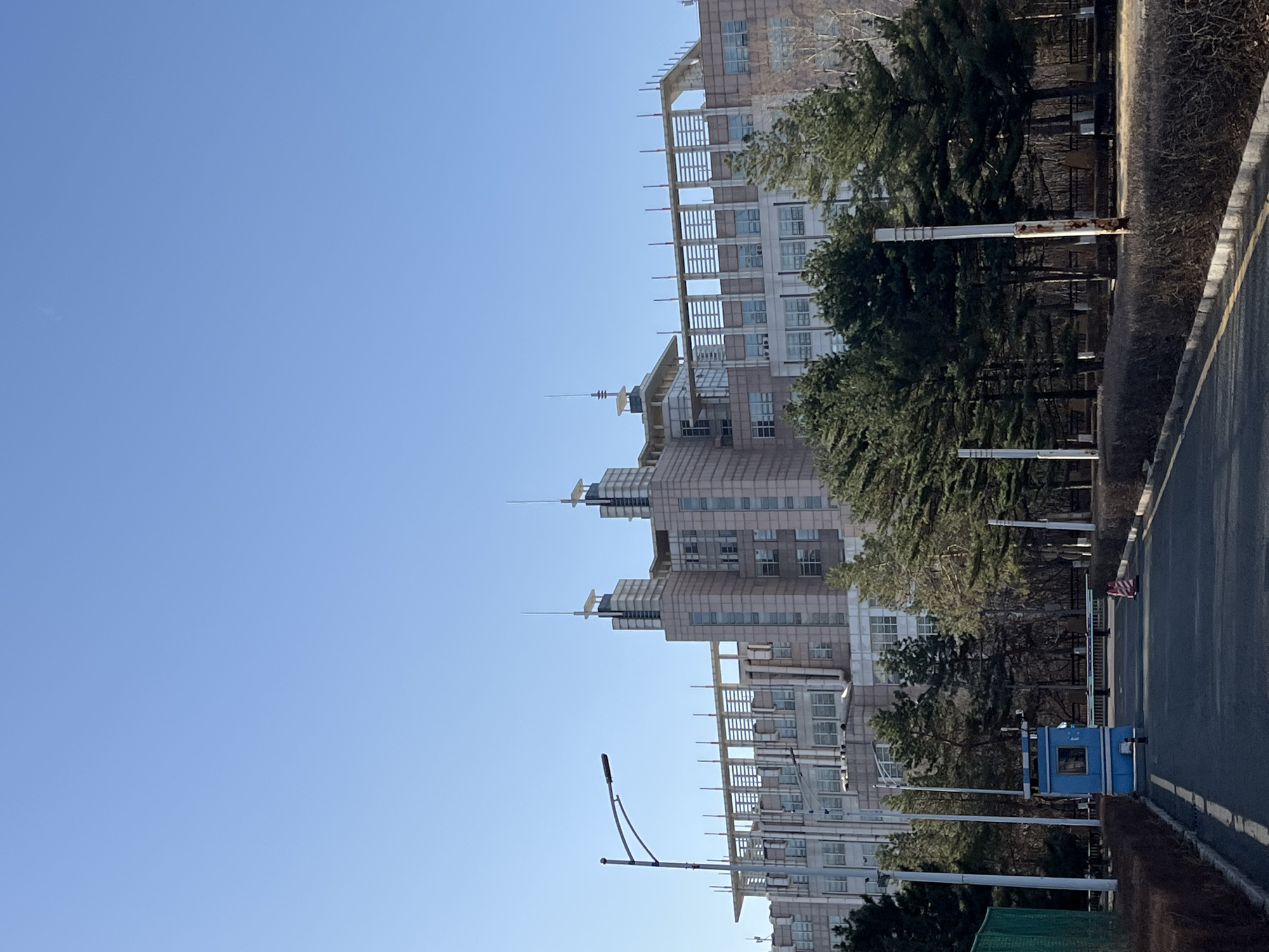 The image depicts an urban landscape in Changchun City, China, showcasing a tall, modern building complex with multiple towers and horizontal architectural features, possibly balconies. The buildings are light in color and appear to be constructed of concrete or similar materials. Adjacent to the buildings, there are mature evergreen trees and roadside vegetation, suggesting a blend of urban development and green spaces. A paved road runs along the right side of the frame, with a blue security booth or utility structure visible near the road. Street lamps are present along the roadway, indicating infrastructure for nighttime illumination. The sky is clear and blue, suggesting a sunny day. No people are directly visible in the frame. The scene appears to be a modern, planned area within Changchun City.