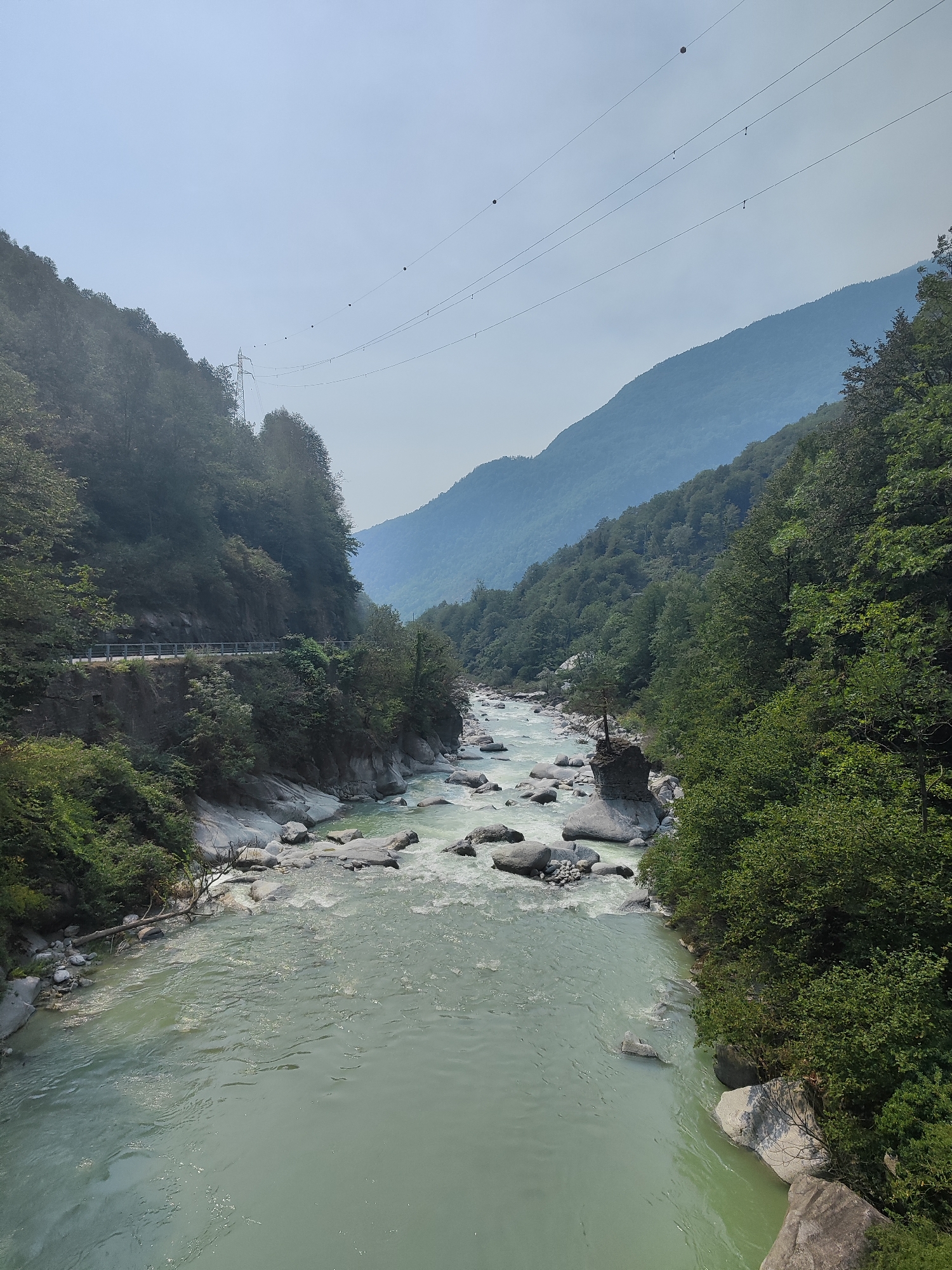 The image depicts a scenic view of the Diveria river flowing through a valley in Varzo, Italy. The river, with its turquoise-colored water, is characterized by rapids and large grey stones. The valley is flanked by steep, tree-covered mountains on both sides. A road with a guardrail is visible along the left side of the valley, built into the rocky slope. In the distance, a network of power lines stretches across the sky, supported by pylons. The sky is hazy and light blue. The overall impression is one of a natural, somewhat wild landscape with evidence of human infrastructure.