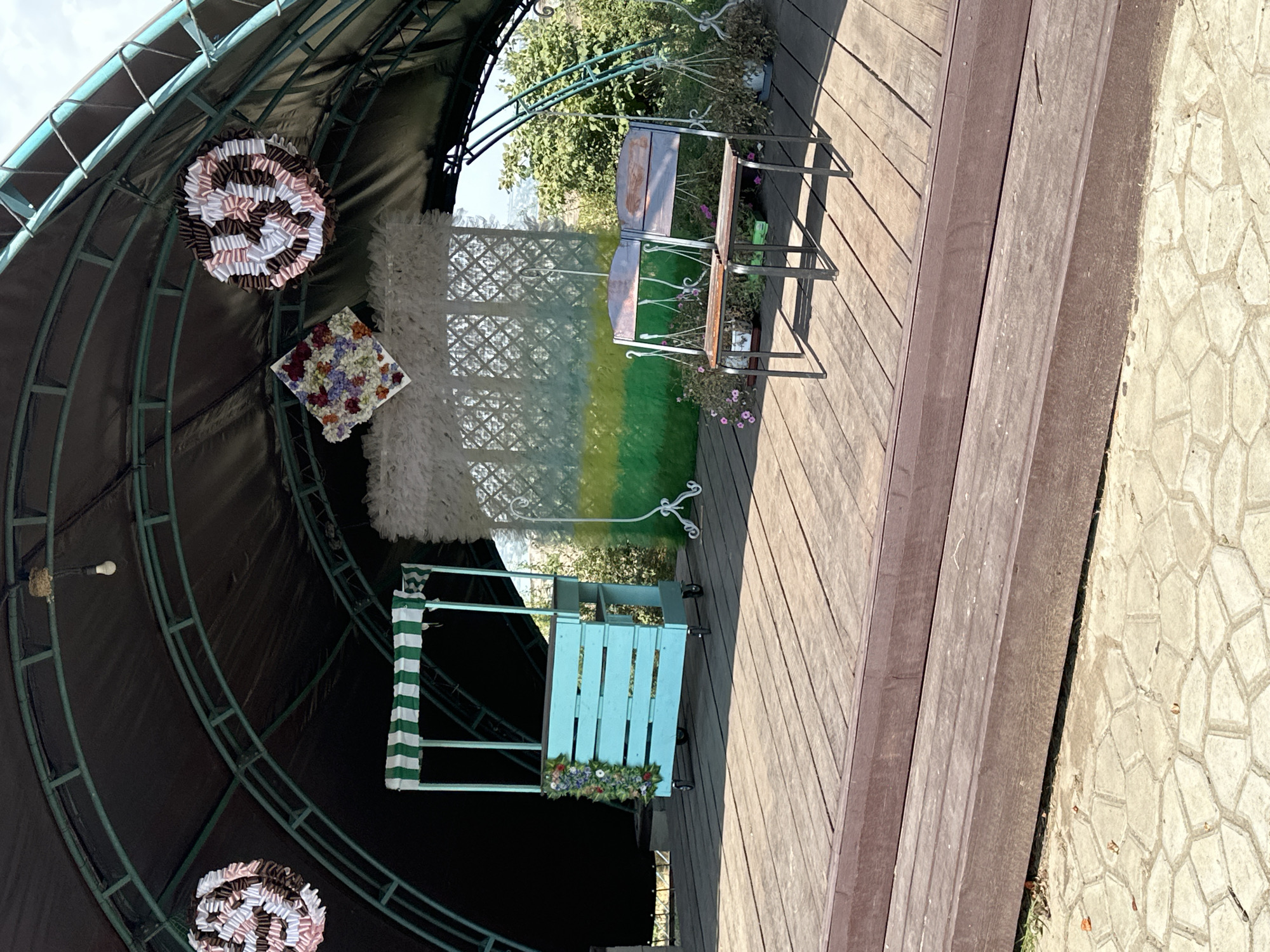 The image depicts an outdoor stage area, likely for events, under a large, arched canopy. The canopy is constructed with dark fabric stretched over a green metal frame. Suspended from the canopy are two circular decorative elements made of folded fabric in shades of pink, white, and brown, and a square floral arrangement.
In the center of the stage is a teal-colored wooden cart with a striped green and white awning. The cart is decorated with a garland of artificial flowers on one side and has wheels for mobility. To the right of the cart, a backdrop appears to be set up, consisting of a white feathery material above a green and yellow painted section, supported by decorative white stands. In front of this backdrop, a two-seater bench made of dark metal and wooden planks is positioned.
The stage itself is made of wooden planks, with a darker wooden step leading down to a paved area with irregularly shaped light-colored stones. The surrounding environment includes greenery and foliage visible behind the stage, suggesting an outdoor setting. The lighting indicates it is daytime, with shadows cast on the stage. The scene is located in the city of Marynivka, Ukraine. There are no people visible in the image.
