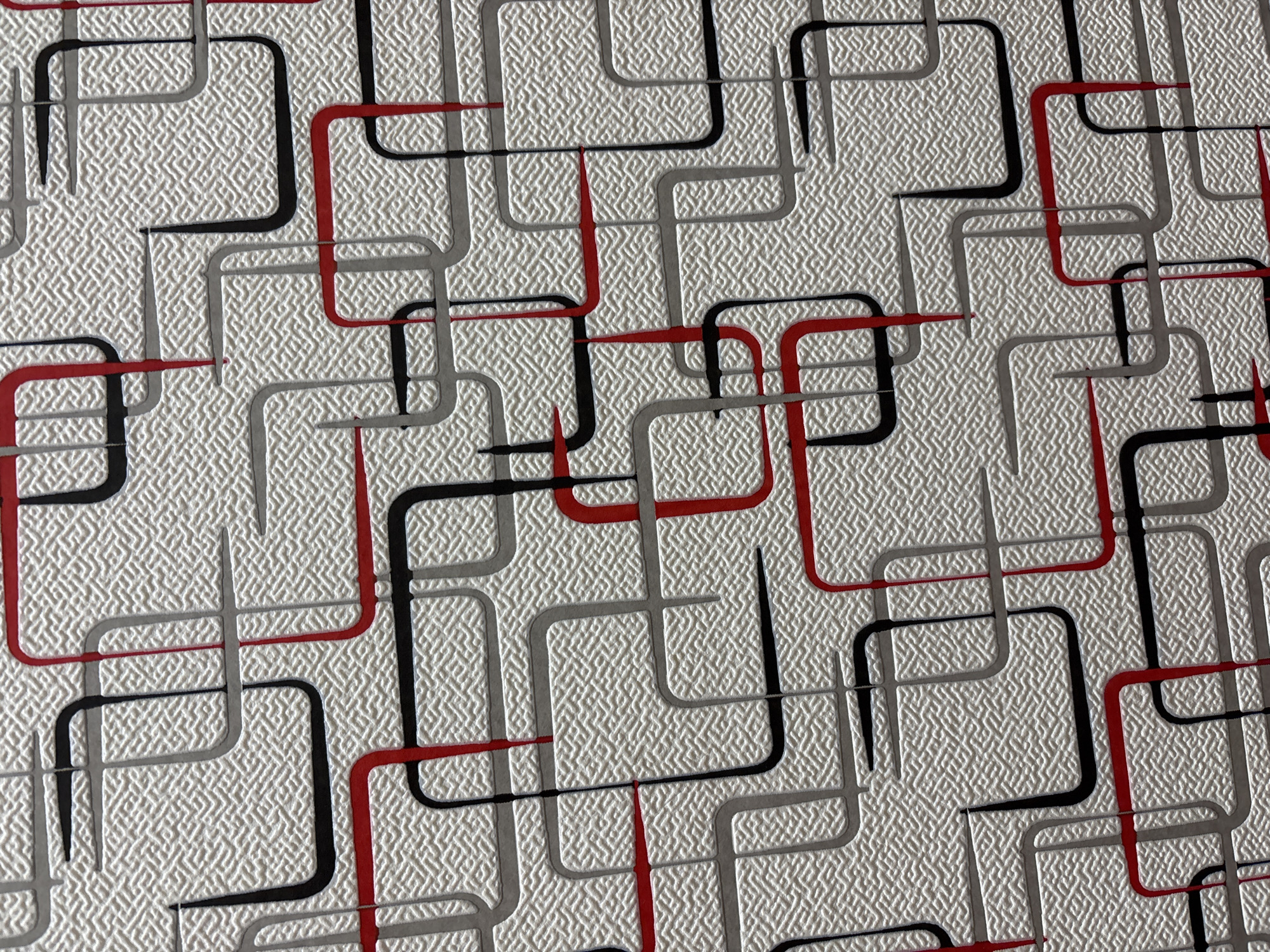 The image shows a close-up, overhead view of a textured wallpaper with a repeating geometric pattern. The pattern consists of interconnected, rounded rectangular shapes in black, red, and gray outlines. The background of the wallpaper has a fine, cross-hatched texture. There are no people, animals, or any discernible environmental features that would indicate a specific location within Novocherkassk, Russia, or any other city. The image solely depicts the wallpaper's surface.