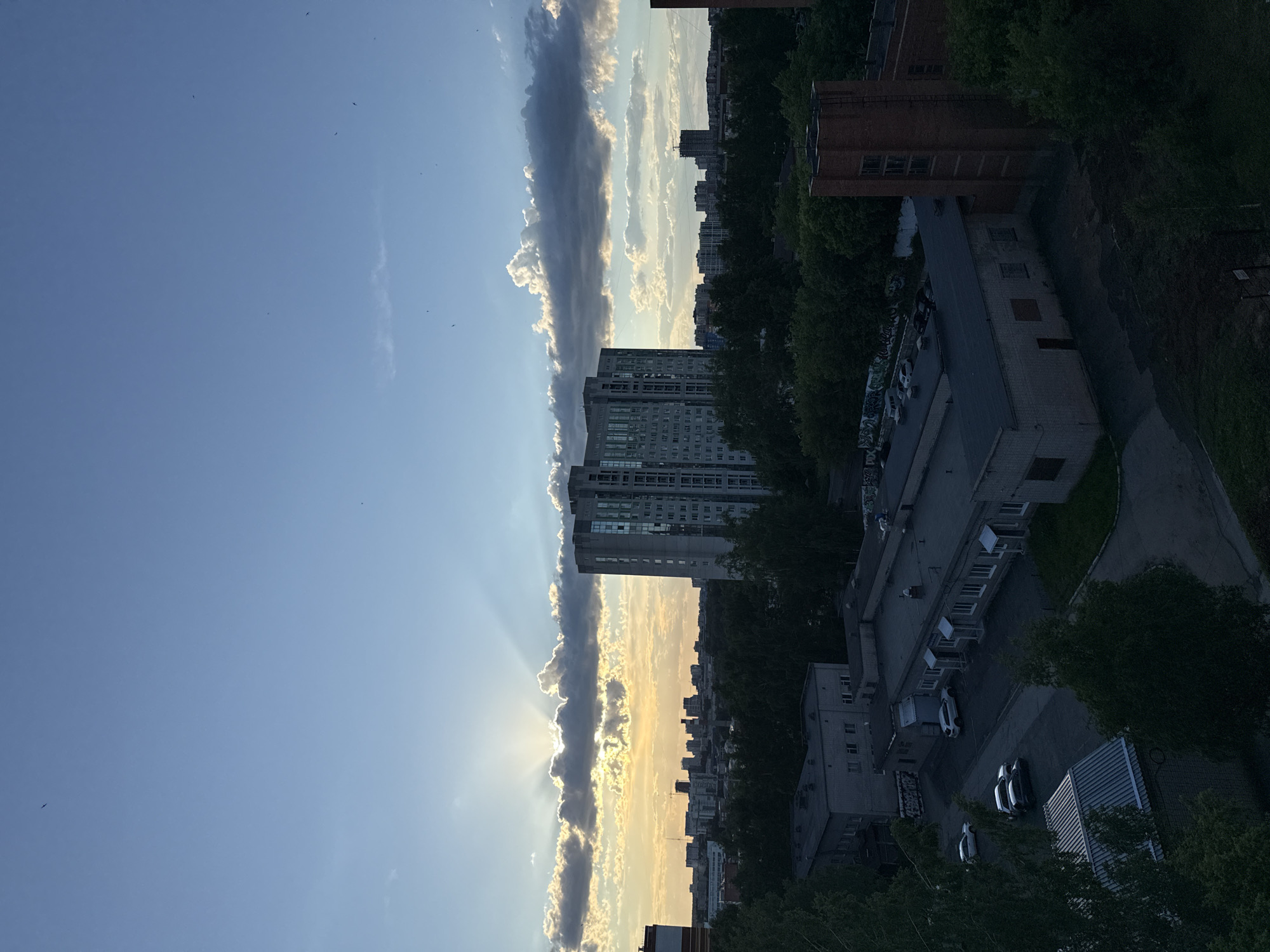 An eye-level view of a cityscape features several buildings, trees, and a partially cloudy sky, with the sun shining behind the clouds and creating rays of light. Tall apartment buildings are visible in the center of the frame, surrounded by lush green trees. To the right, lower-rise buildings and parking lots can be seen, while the left side of the frame is dominated by the blue sky with clouds. The location is Izhevsk, Russia.