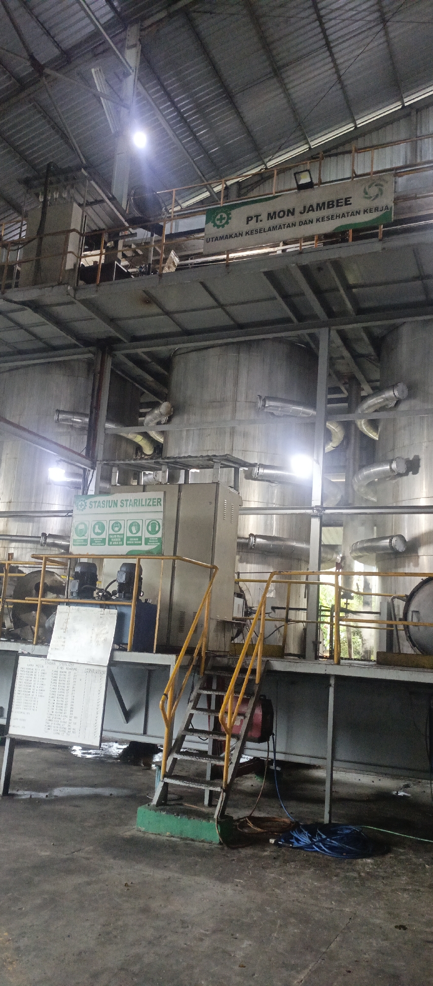 The image depicts the interior of an industrial facility, likely a factory or processing plant, in the city of Iemirah, Indonesia. The scene is dominated by large cylindrical tanks and an intricate network of pipes and machinery. A prominent green and white sign reads "PT. MON JAMBEE" and "UTAMAKAN KESELAMATAN DAN KESEHATAN KERJA," which translates to "Prioritize Safety and Health at Work." Another sign indicates "STASIUN STABILIZER" (Stabilizer Station) and displays several safety icons, including one for eye protection.
The facility appears to be functional, with visible equipment like electric motors mounted on a platform. A metal staircase with yellow railings leads up to this platform. In the foreground, a concrete floor shows signs of wear and some dampness, with electrical cables and hoses coiled on the ground. The overall environment suggests a working industrial setting, with ample lighting from overhead fixtures and natural light filtering in from unseen sources. No individuals are visible in the image.