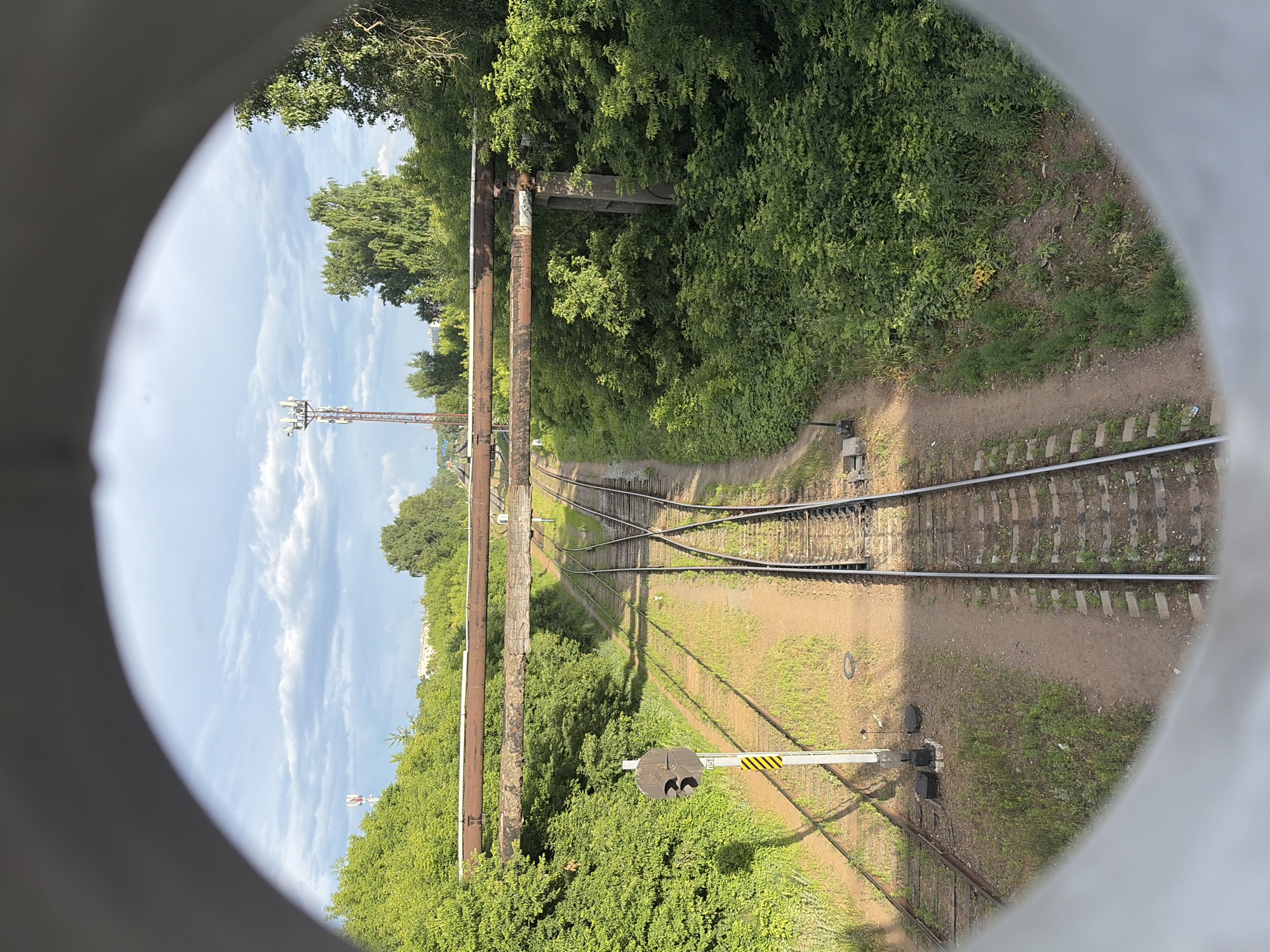 This media file is an image looking through a circular frame at a railway track junction in Chaikovsky, Russia. The scene depicts a sunny day with a partly cloudy sky. Lush green trees and vegetation line the sides of the railway, creating a natural environment around the industrial infrastructure.
The main subject is the railway track system, with multiple sets of tracks converging and diverging, indicating a junction. In the distance, a tall telecommunications tower is visible against the sky, suggesting the presence of modern infrastructure in the vicinity. A railway signal, with a yellow and black striped arm, stands near the tracks, indicating its function in controlling train movement. There are also two large, rusty pipes running parallel to the tracks, possibly part of an overhead bridge or industrial pipeline.
The image appears to be taken from an elevated position, looking down at the tracks. The circular frame through which the scene is viewed suggests the vantage point might be through a pipe, a tunnel, or a similar circular opening. The lighting and shadows suggest it is daytime, likely late morning or early afternoon. The weather appears to be pleasant and clear, suitable for outdoor activities. No people are visible in the image, and the overall mood is one of observation of the landscape and infrastructure.