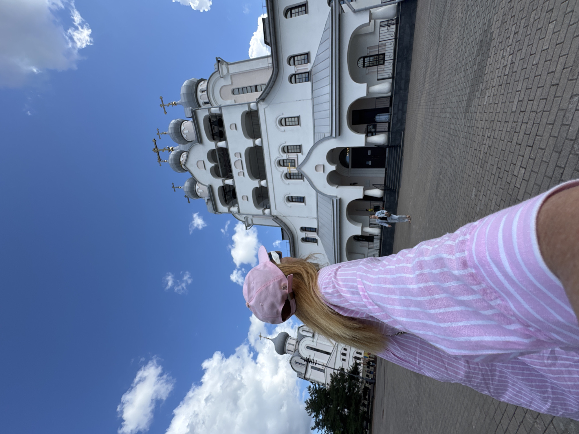 A blonde woman wearing a pink baseball cap and a pink and white striped shirt stands in the foreground, facing away from the camera and looking towards a white church with multiple gray domes and crosses. The church has arched windows and multiple levels, with a bell tower on the left side. The ground in front of the church is paved with gray bricks. The sky is blue with scattered white clouds. Another person in a blue shirt and jeans is walking in the background towards the church. The scene is likely a religious site in Saint Petersburg, Russia, given the architecture and the presence of a church with onion domes, a common feature of Russian Orthodox churches.