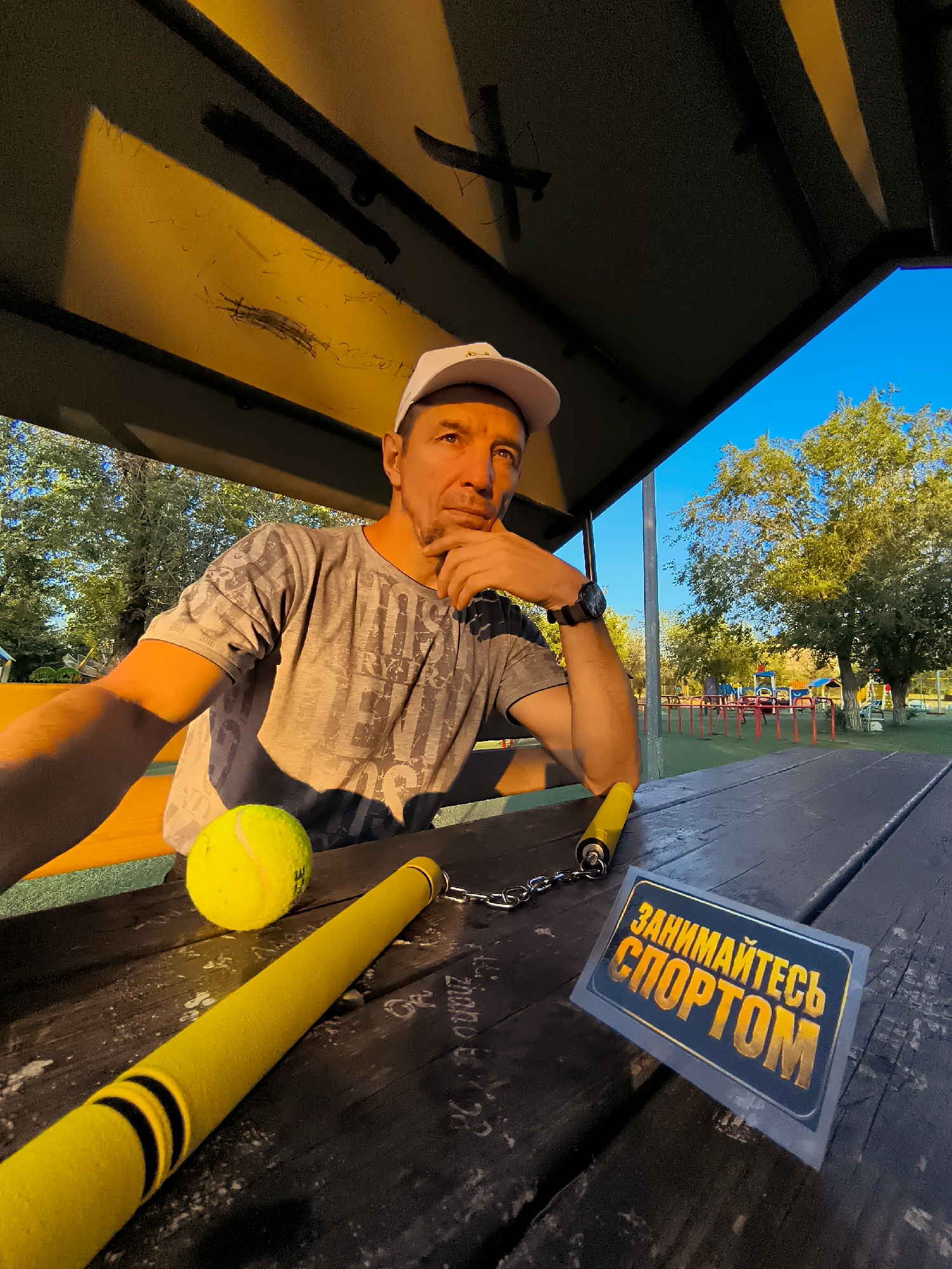 A man wearing a white baseball cap and a grey t-shirt sits at a wooden picnic table. He is resting his chin on his hand and looking towards the camera. On the table in front of him are a yellow tennis ball and a yellow nunchaku. To the right of the nunchaku is a sign with Russian text that reads "ЗАНИМАЙТЕСЬ СПОРТОМ" which translates to "DO SPORTS". The table is set outdoors, with a playground visible in the background. The scene appears to be in Qarağandı city administration, Kazakhstan.