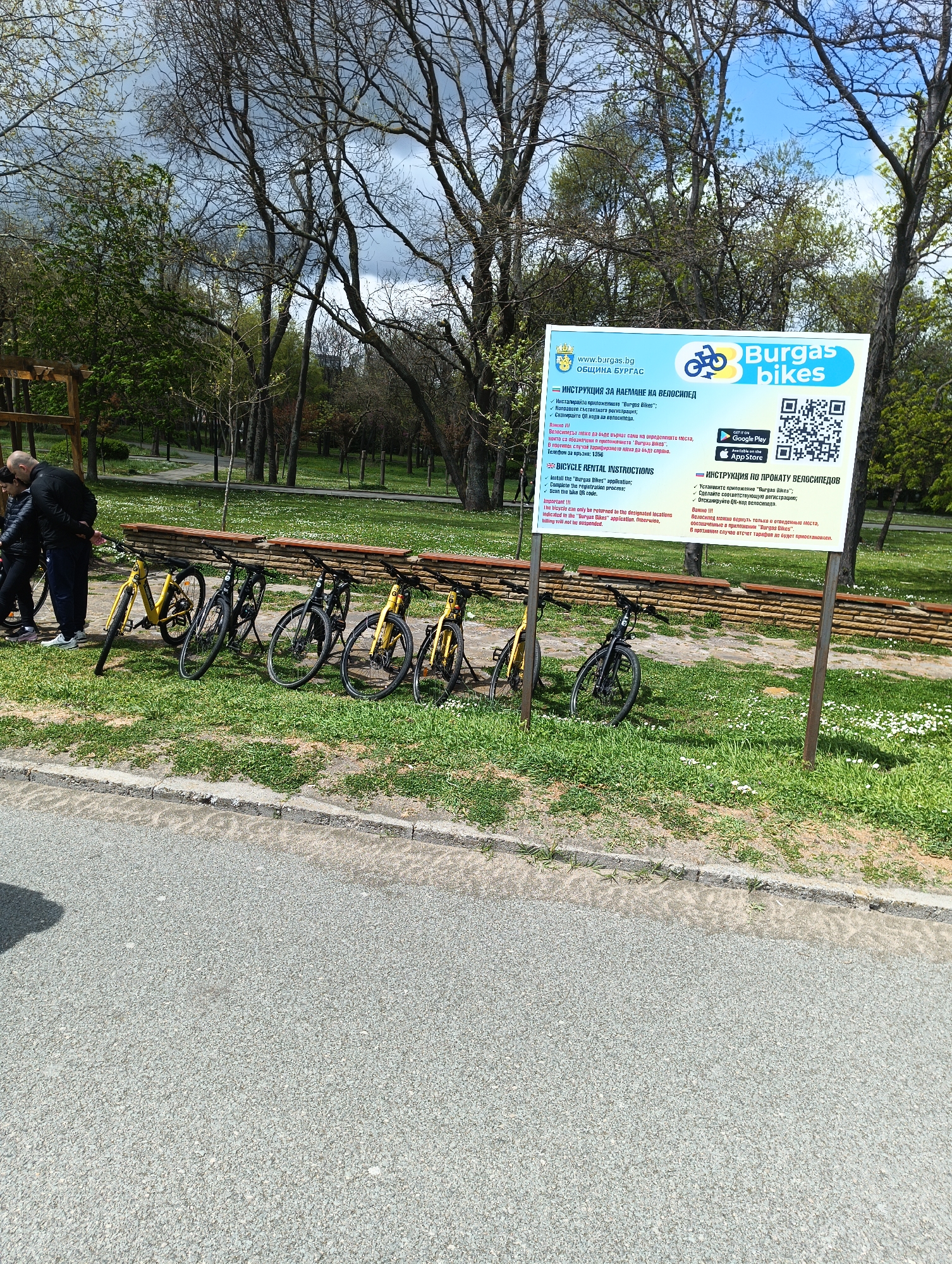 The image depicts a scene in Burgas, Bulgaria, featuring a row of public bicycles provided by "Burgas Bikes". The bikes, a mix of black and yellow, are parked alongside a low brick-and-wood divider in a park setting. To the left, two individuals, presumably intending to rent or having just rented a bike, stand near the bicycles. Behind them is a lush park with trees and green spaces.
To the right of the bikes is a large sign outlining instructions for renting the bicycles in Bulgarian, English, and Russian. The sign also features the "Burgas Bikes" logo, app download information, and a QR code. The background shows more trees and greenery, suggesting a park or recreational area.
In the foreground, a paved path leads to the scene, marked by a concrete curb separating it from the grassy area. The weather appears overcast with hints of sunlight peeking through the trees.