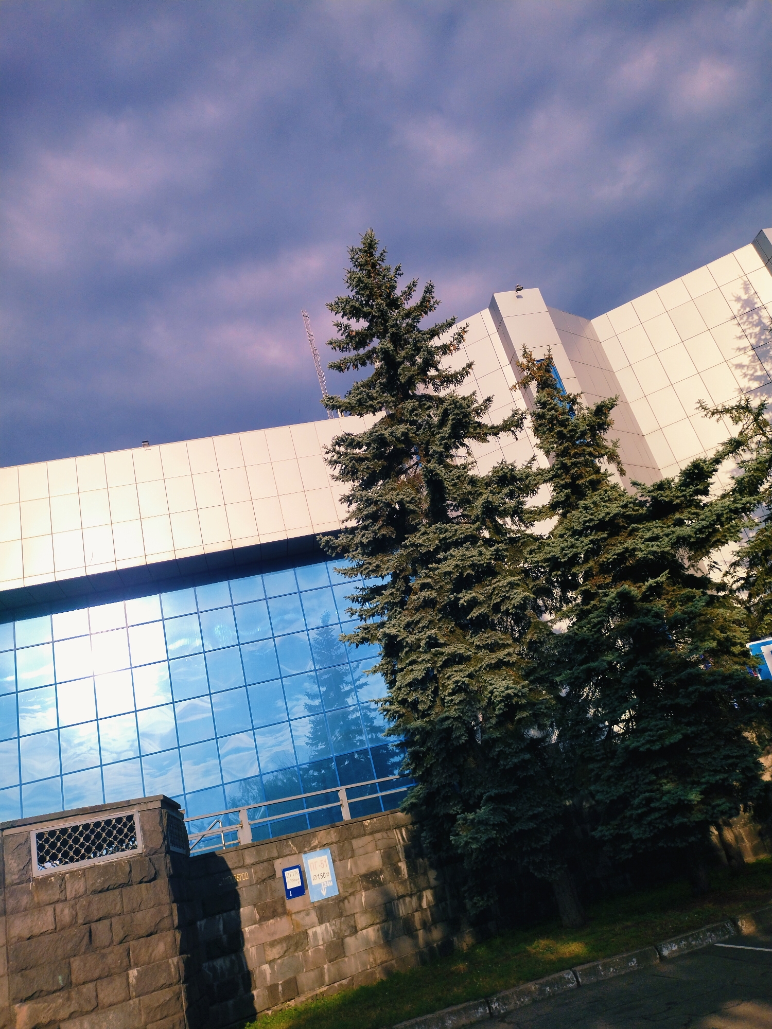 The image depicts a modern building with a facade of white panels and a large section of blue glass windows, reflecting the sky and surrounding trees. In front of the building, there are several mature evergreen trees, their branches reaching upwards towards the building and the cloudy sky. A stone wall with metal grates and signage is visible in the foreground, bordering a paved area that appears to be a road or walkway. The scene is set in the city of Horishni Plavni, Ukraine.
