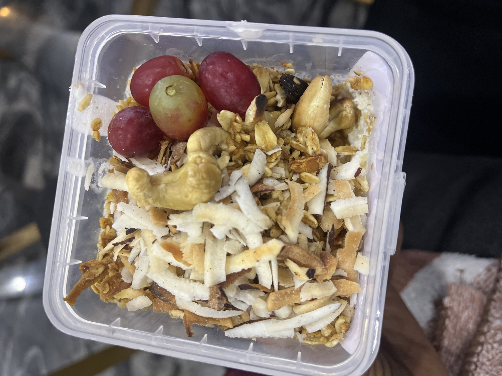 The image displays a clear, rectangular plastic container, held by a person's left hand, against a blurred, dark background. The container is filled with a layered food item. The bottom layer consists of a light purple or white creamy substance, likely yogurt. Above this, a mixture of granola or toasted cereal forms a base. Toasted coconut flakes, varying in color from white to light brown, are prominently scattered over the granola. Several whole cashews are visible, along with some other nuts, possibly peanuts, and a dark, dried fruit such as a raisin. Positioned towards the upper right of the container are three grapes: two red and one green, with a small stem fragment attached to the green grape. The hand holding the container is partially visible, showing skin tone and a portion of a soft, patterned garment featuring pink and white colors. The background is out of focus, appearing as dark, undefined surfaces with faint reflections of light sources. This scene is observed in the city of Abuja, Nigeria.