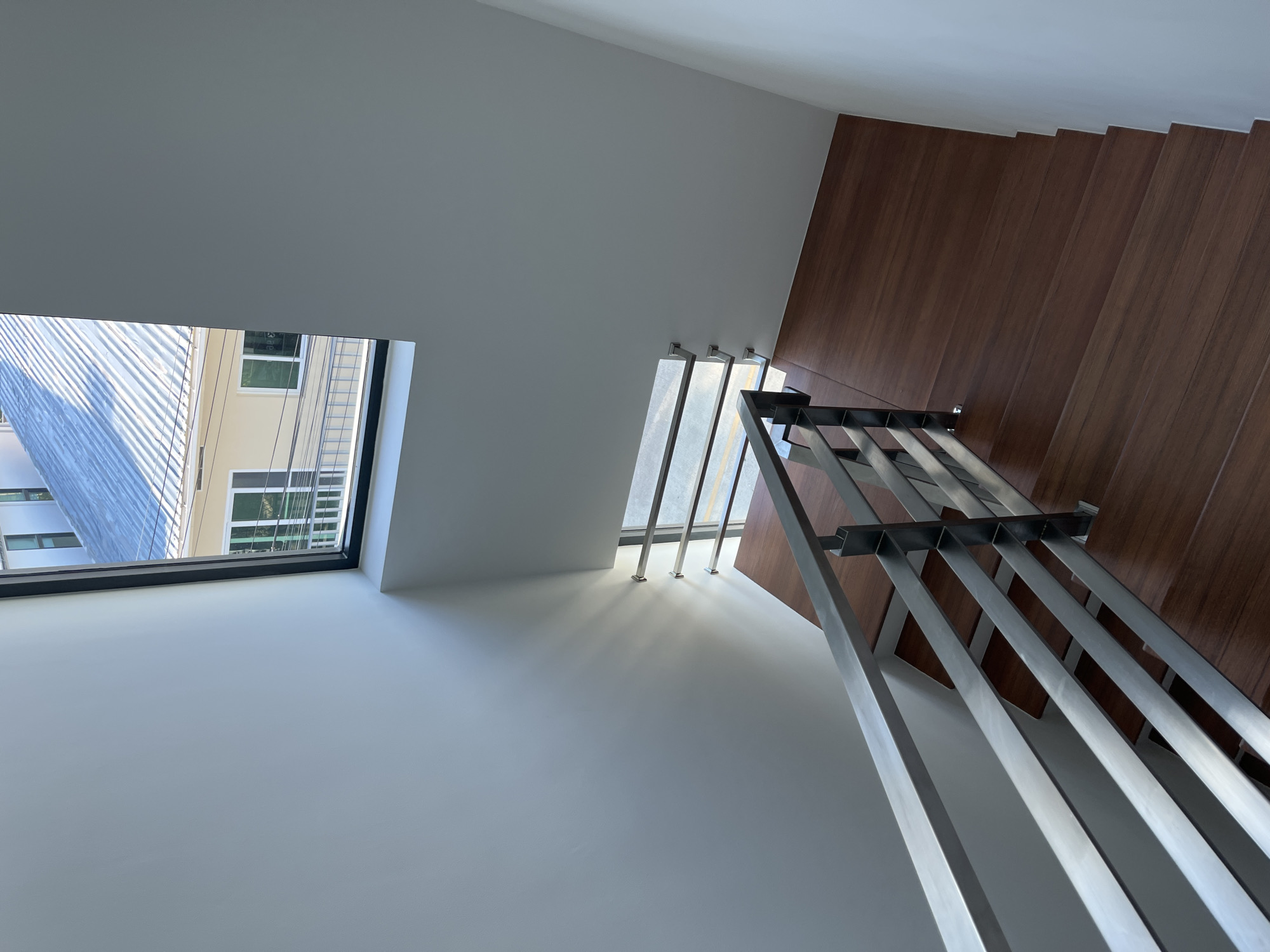 An interior view looks down a modern staircase with dark wood treads and a sleek stainless steel railing. The staircase is illuminated by natural light streaming through a large, high window, casting shadows on the white wall. Outside the window, a corrugated metal roof of a building is visible, along with some power lines and a portion of another building with a balcony. This scene is located in a residential building in Rawai, Thailand.