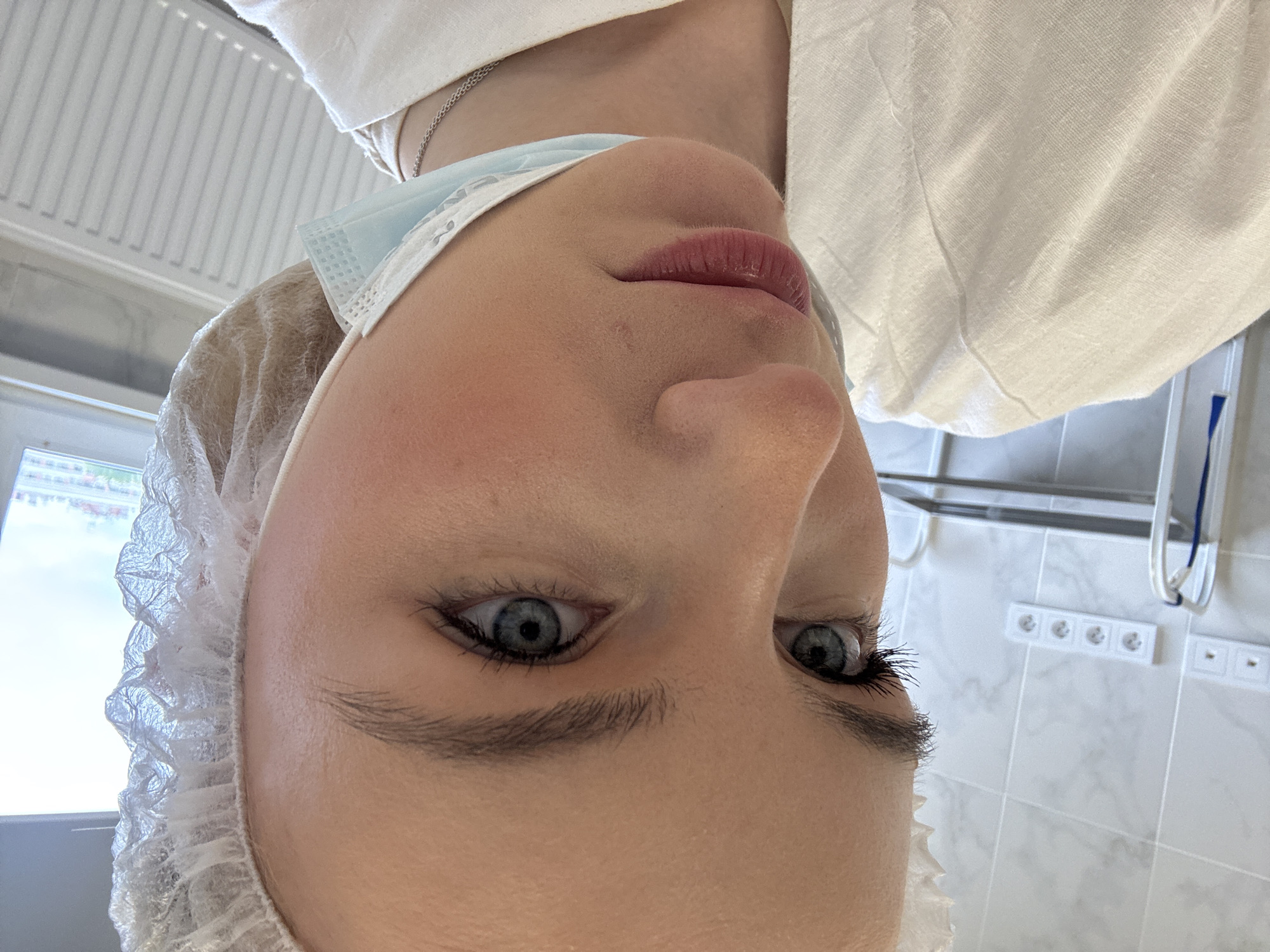 A close-up, inverted view shows a woman in medical attire. She wears a light blue surgical mask pulled down below her nose, a white bouffant cap, and a white top. The background features a tiled wall with electrical outlets and a radiator, suggesting an institutional setting. The location may be Saint Petersburg, Russia.