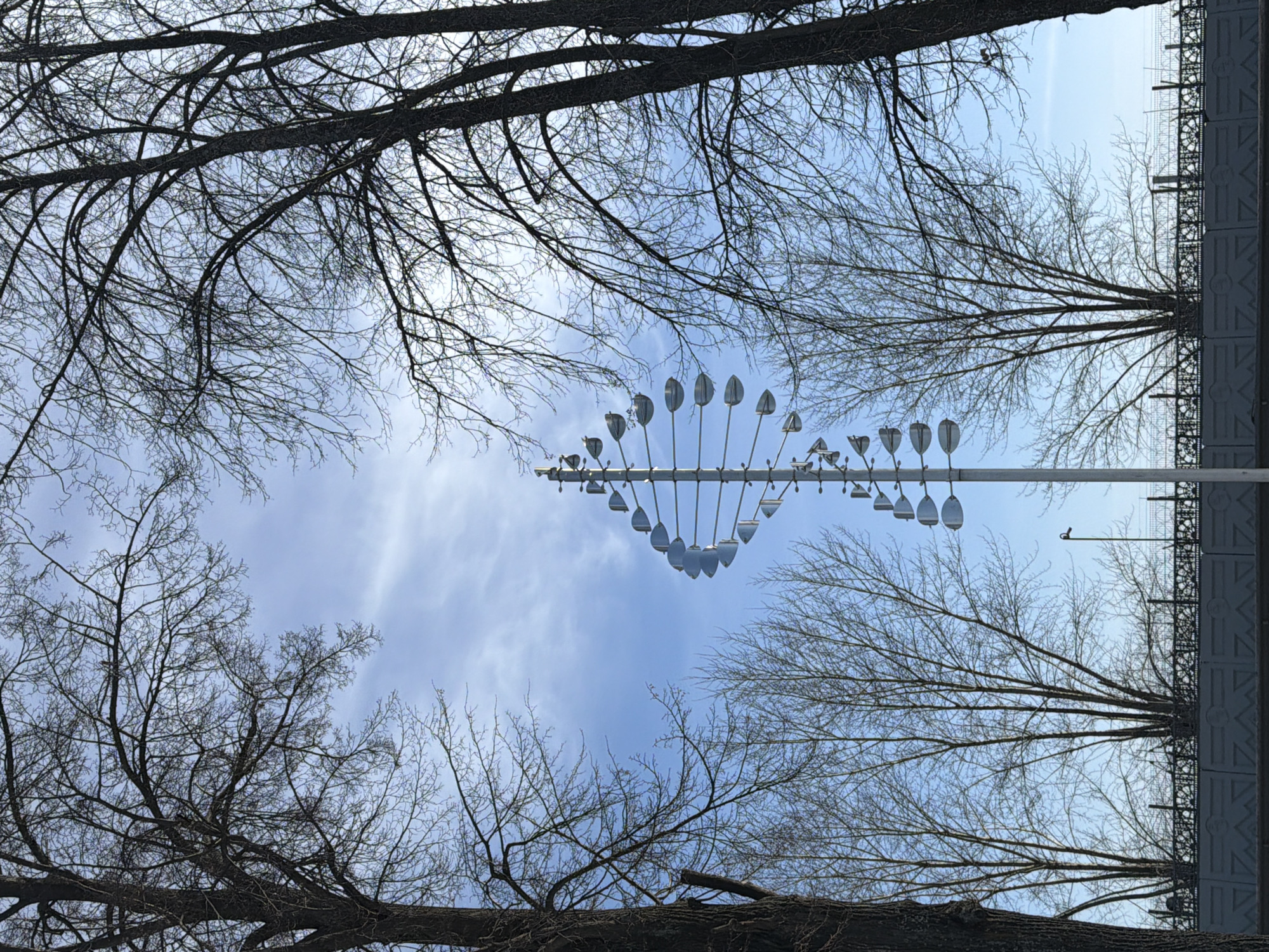 Captured from below in Votkinsk, Russia, a sky view is framed by bare tree branches in winter. An artistic wind chime structure resembling a DNA helix is suspended in the center, featuring reflective teardrop-shaped elements. In the background, a cloudy blue sky suggests overcast weather. To the right, a modern architectural structure with a repeating design is visible. No people are present.