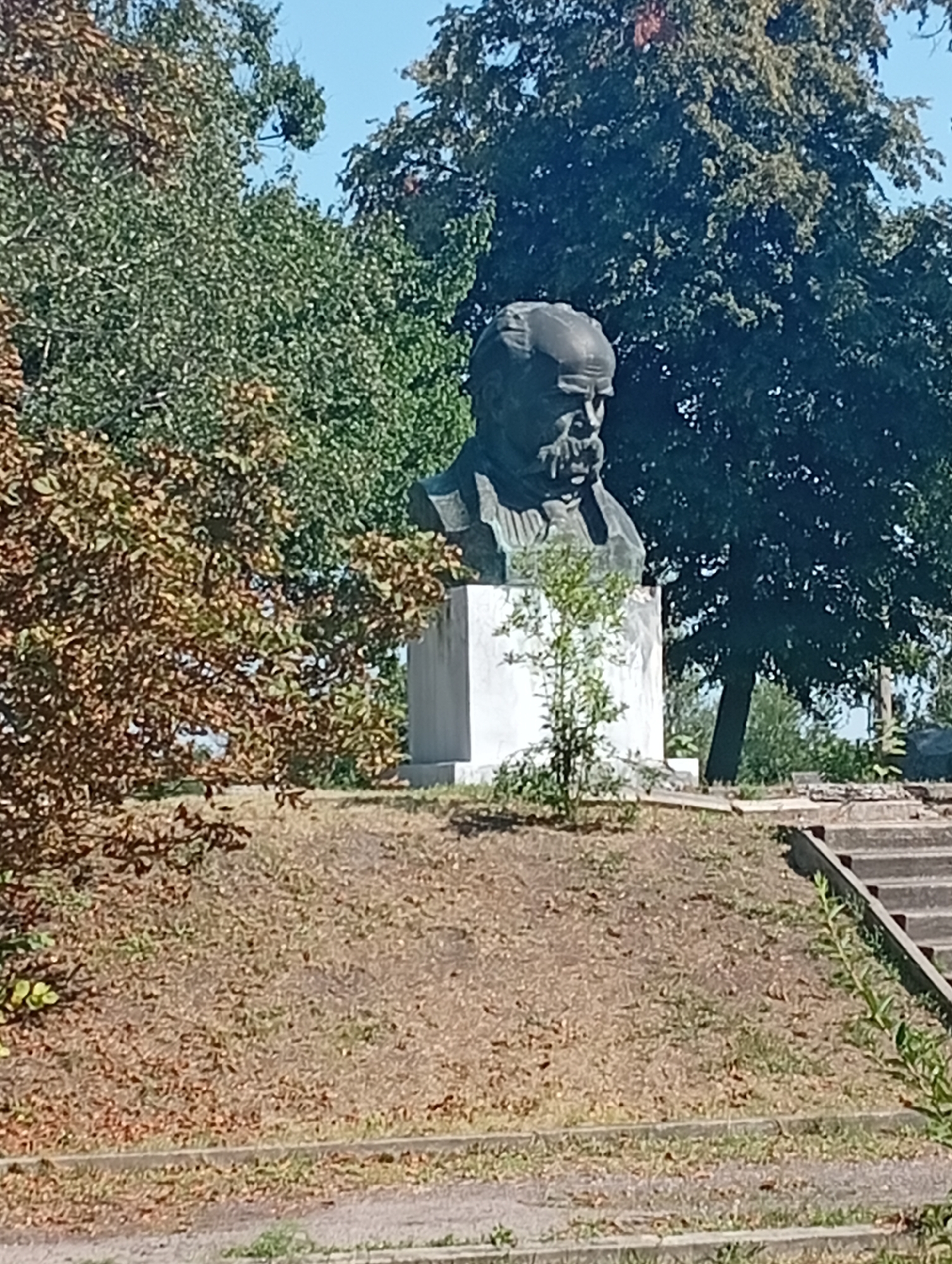 The image shows a bust of a man on a white pedestal in Khomutets, Ukraine. The bust is made of dark metal and depicts a man with a beard and mustache, wearing a hat. The pedestal is square and sits on a grassy hill. Behind the bust, there are trees and a clear blue sky. In the foreground, there are some steps leading up to the pedestal. The overall scene is peaceful and serene.