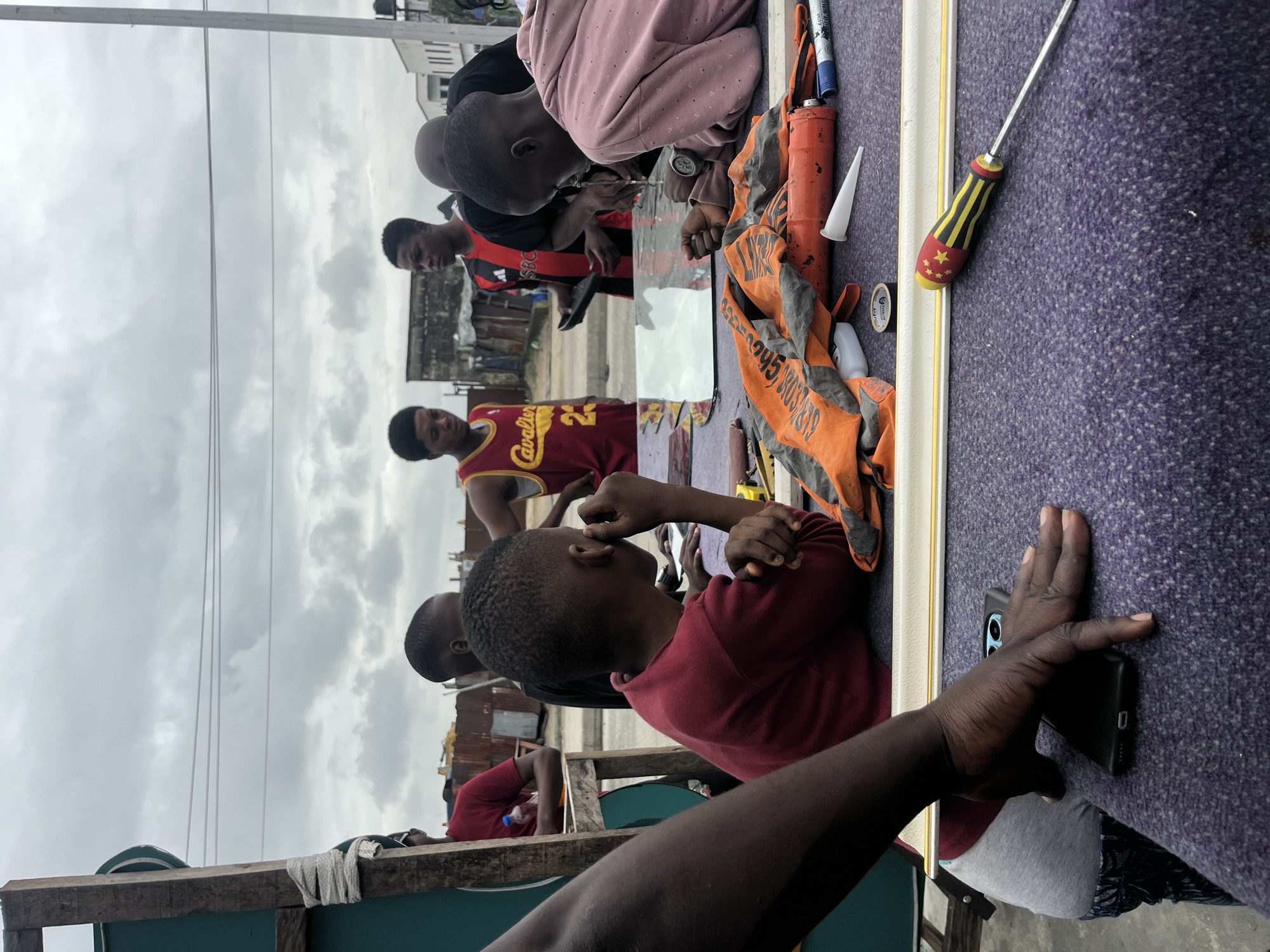 In Ado, Nigeria, a group of young men are gathered around a table outdoors, seemingly engaged in a craft or repair project. The table is covered with a purple textile and displays various tools, including a screwdriver with a red and yellow striped handle, a caulking gun, rolls of tape, and a small tube of adhesive. A framed mirror lies on the table, suggesting they might be working on framing or repairing it. Several of the young men are wearing casual clothing, including a basketball jersey with "Cavaliers 23" on it. One person in a pink hooded jacket is leaning over the table, appearing to focus intently on the task. The background reveals a somewhat weathered urban environment with corrugated metal structures and buildings, under an overcast sky. The overall scene depicts a collaborative effort in a practical, everyday setting.
