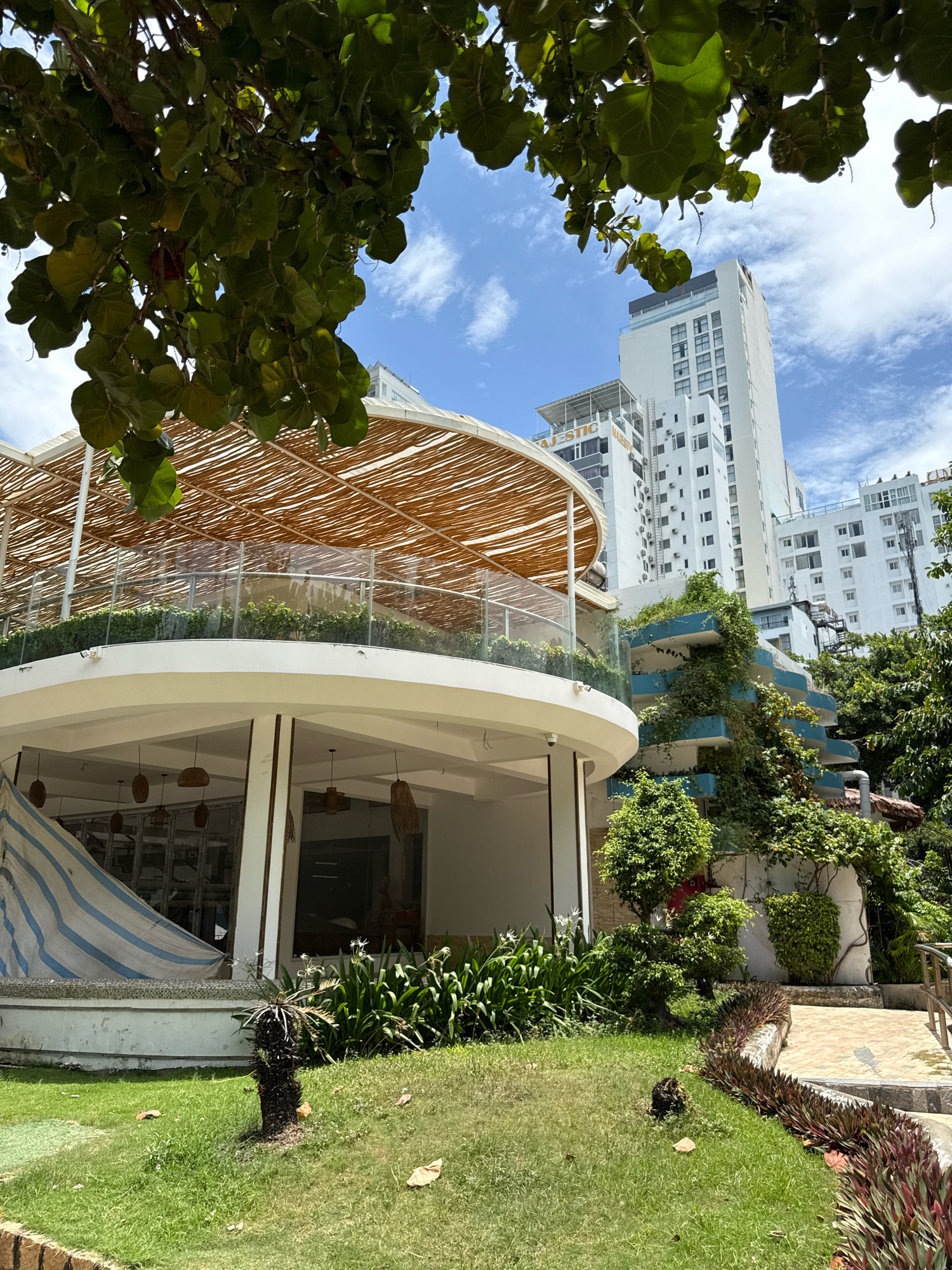 The image shows a building in Nha Trang, Vietnam. The building is white and has many windows. There are trees and plants around the building. The sky is blue with some clouds. There is a circular balcony on the building with a thatched roof. There are also some hanging lamps visible. In the foreground, there is a patch of grass with some fallen leaves. To the right, there is a pathway leading up to the building.
