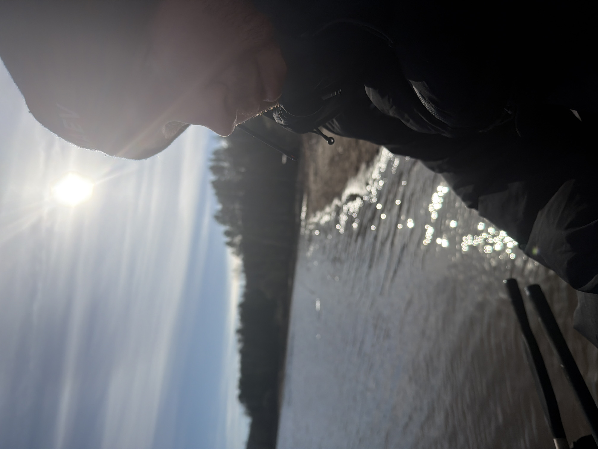 The image, originally rotated 90 degrees, depicts a male figure in a backlit outdoor setting, likely near Chaikovsky, Russia. The man is seen in profile on the right, looking left towards a sun-drenched body of water. He wears a dark jacket and a light-colored beanie with faint text "NEV" or "ΛEV" on it. His face is largely in shadow due to the intense backlighting from the sun, which occupies the upper left, casting strong rays and lens flare.
The body of water, possibly a river or lake, dominates the foreground and midground, characterized by sparkling, reflective surface patterns from the bright sun. A dense line of dark evergreen trees forms a forest along the opposite bank, silhouetted against the bright, slightly cloudy sky. Partially visible in the lower right are two dark poles, potentially ski or fishing rods, indicating an outdoor activity. The scene suggests a clear, sunny day, with the person engaged in observation or leisure by the water.