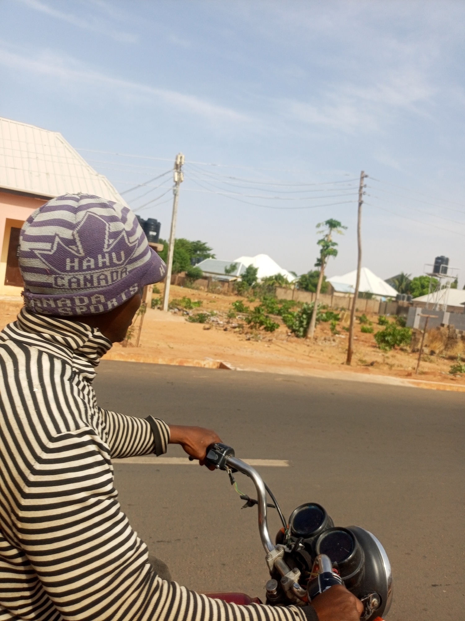 A man wearing a striped turtleneck sweater and a hat is riding a motorcycle down a paved road in a suburban area of Unknown, Unknown. The man is facing away from the viewer and toward the front of the motorcycle, with his hands on the handlebars. In the background, there are houses with white roofs, trees, and utility poles. The sky is blue with light clouds. The environment appears to be dry and dusty.