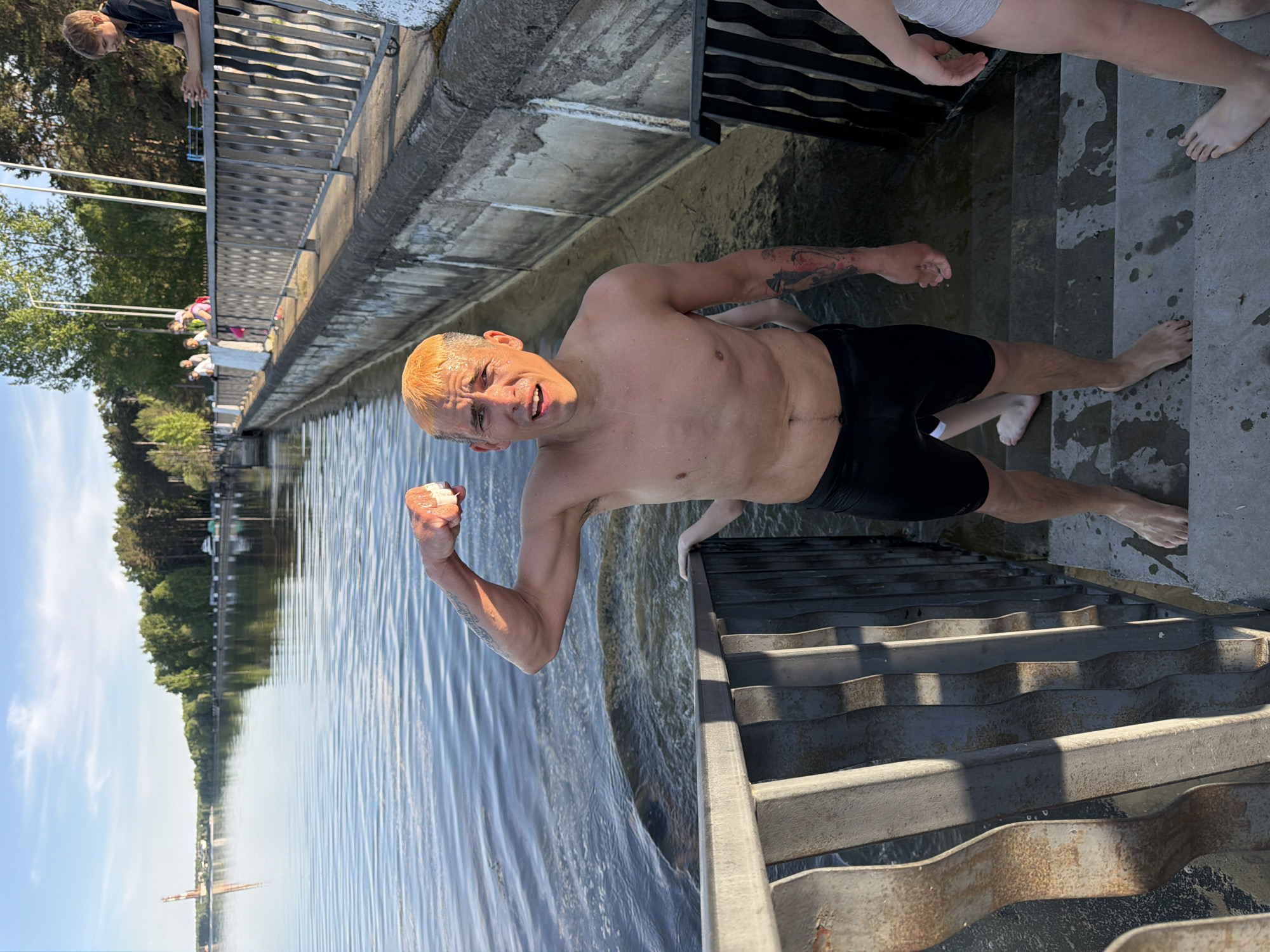 A man in black swimming trunks stands waist-deep in water alongside a concrete pier, possibly a public bathing area. He is holding a white object in his raised right fist, and his left hand appears to grip the pier's steps for support. The steps are made of metal and lead directly into the water. Additional people, including a boy looking over a railing, are on the pier near the steps. The water has ripples, and trees and buildings are visible in the distance. The location is the city of Chaikovsky, Russia.