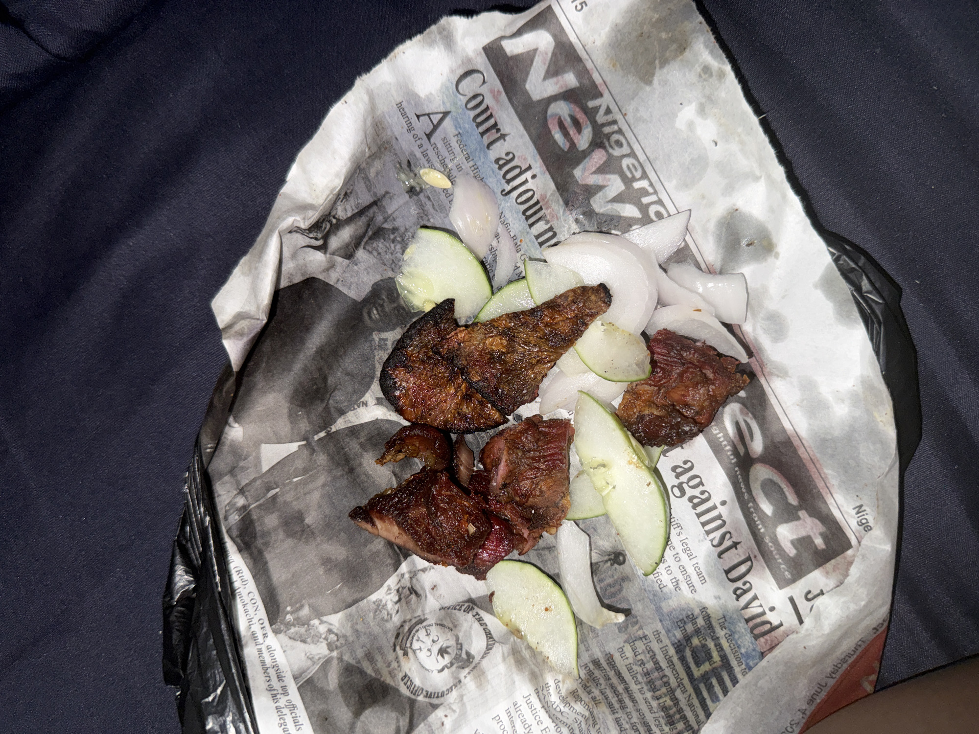 The image shows a serving of grilled meat with sliced onions and cucumber on top of a newspaper. The newspaper appears to be the "Nigerian Tribune" and the headline "Court adjourn" is visible. The food is wrapped in a white paper, and a black plastic bag is also present in the image. The setting is dark and appears to be indoors, possibly a meal being consumed in a casual setting in Abuja, Nigeria.