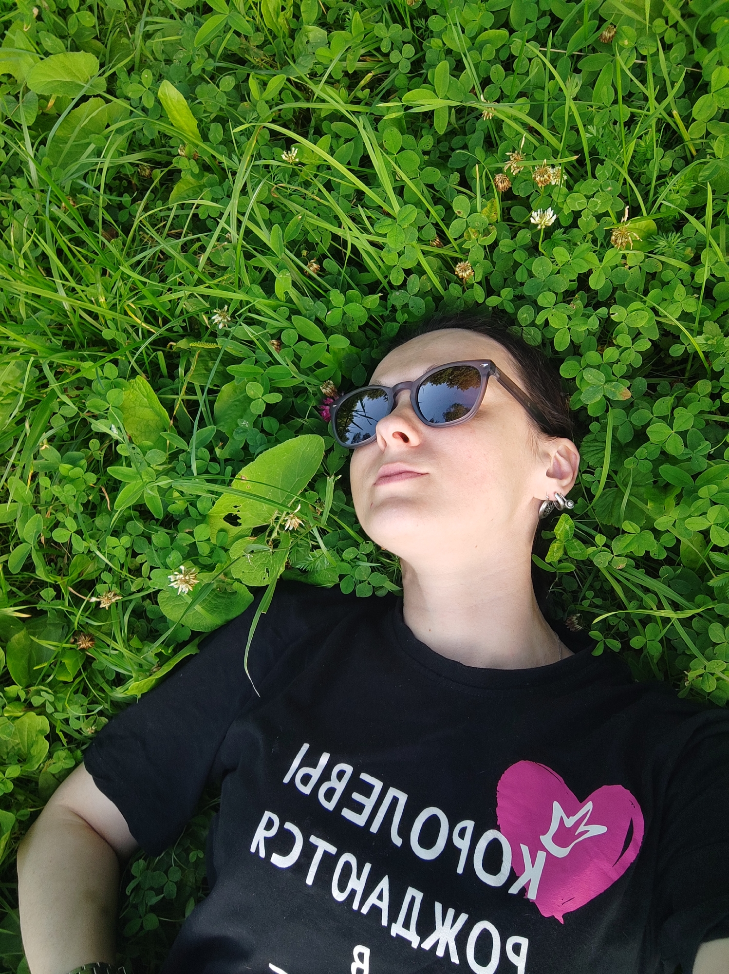 A woman lies on her back in a field of green clover and grass, looking up and to the right. She is wearing a black t-shirt with white text and a pink heart with a crown inside it, sunglasses with dark lenses, and silver hoop earrings. The scene is outdoors, possibly in a park or field in the city of Alabino, Russia, as indicated by the surrounding clover and grass. The lighting suggests it is daytime, likely with sunlight filtering through the foliage.