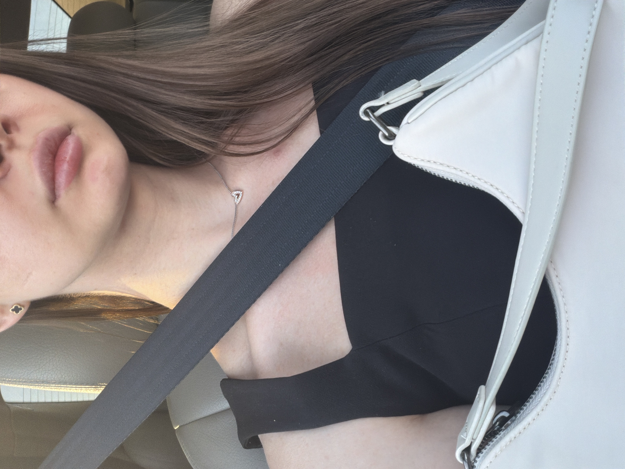 A person is seen from the chest up, wearing a black dress and a seatbelt. They are in a car, indicated by the leather seat and the seatbelt. A white purse is visible in the foreground. The person has long brown hair and is wearing a necklace with a heart-shaped pendant and earrings. The lighting suggests it is daytime.